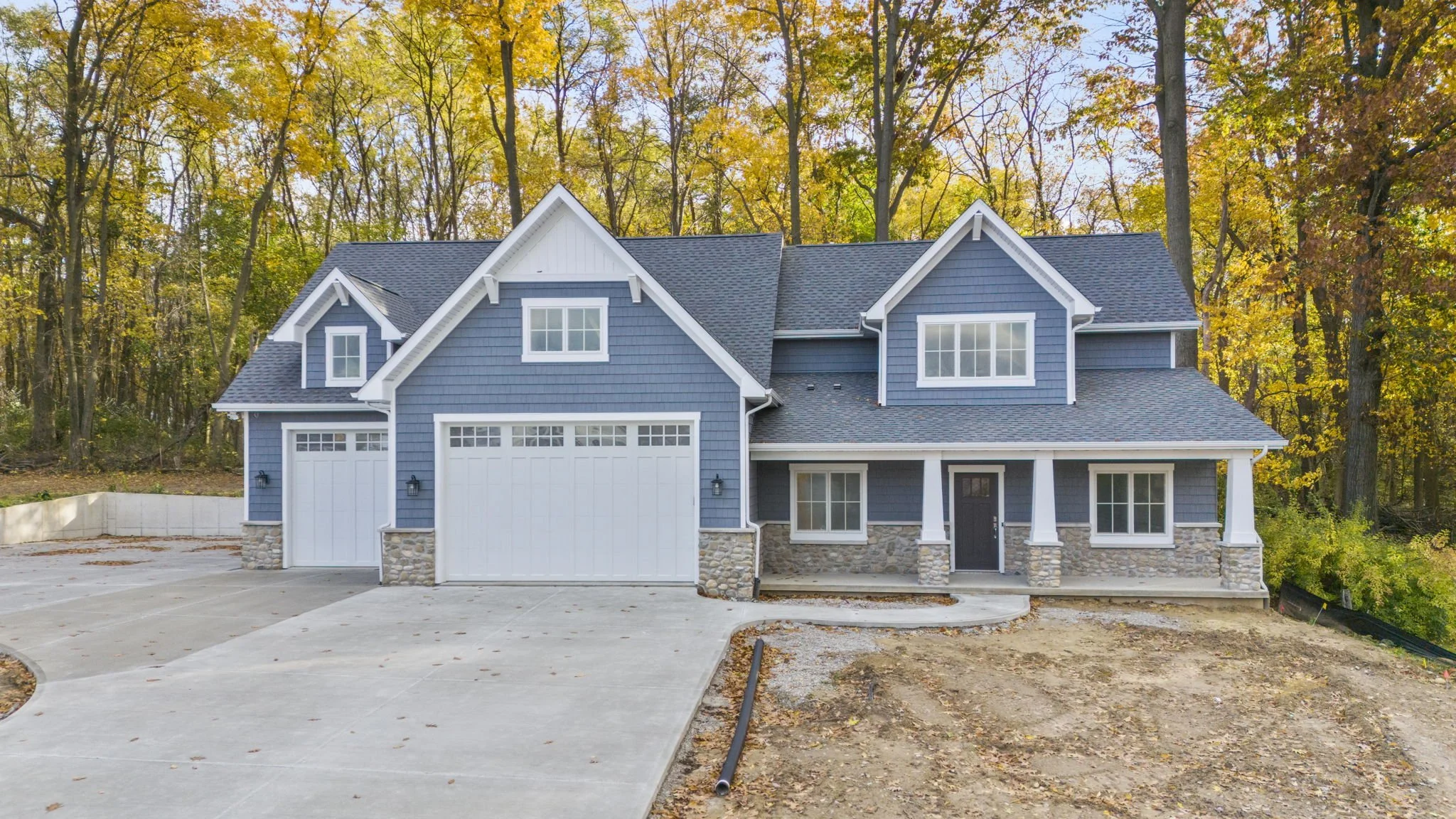 New two-story blue house with white trim, stone accents, and a dark roof, situated in a wooded area during fall with yellow and orange leaves.