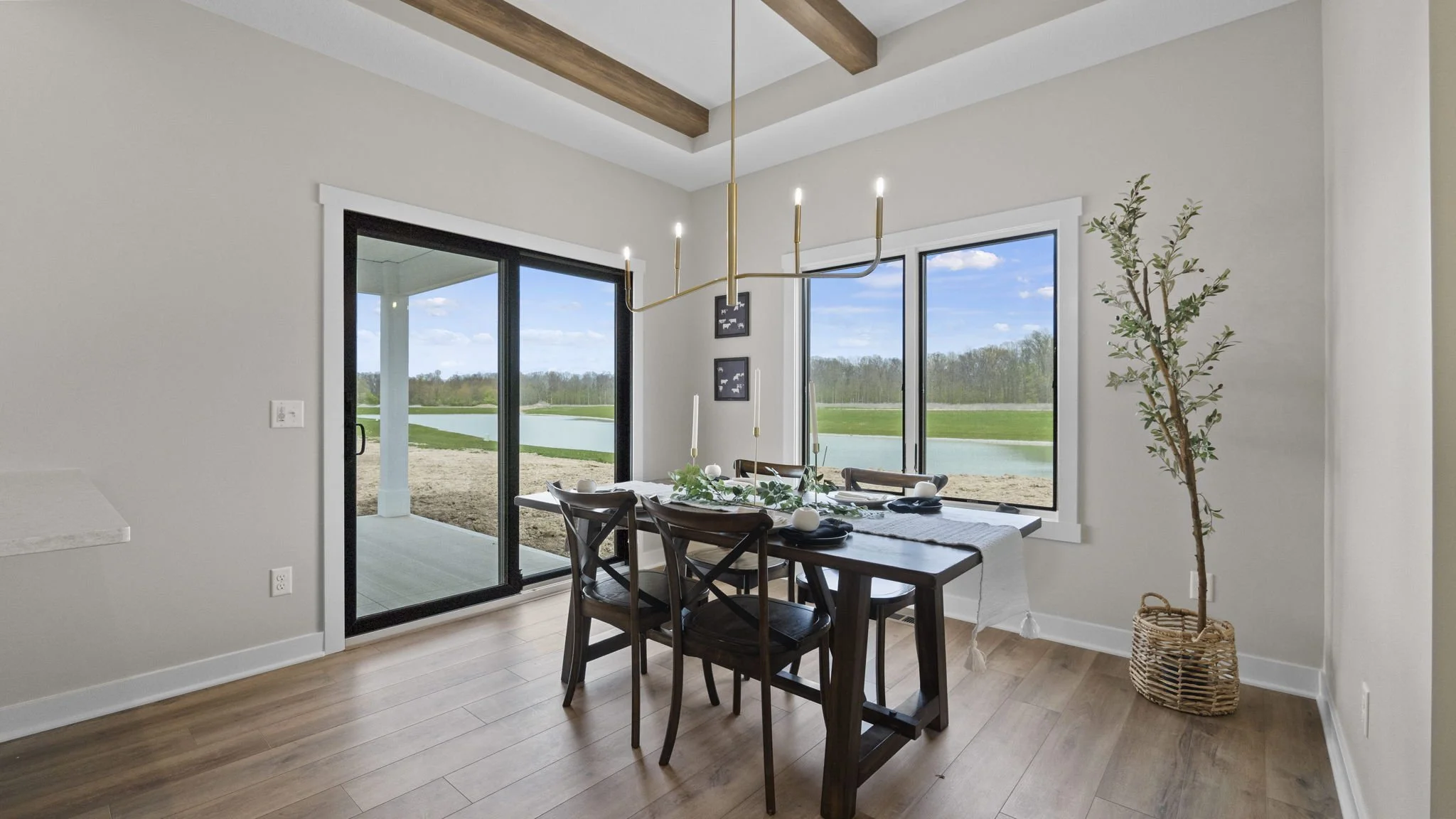 Dining room with a wooden table and chairs, decorated with candles and greenery, next to large windows and sliding glass door overlooking a lake and outdoor space, with a ceiling chandelier and a potted tree in a wicker basket.
