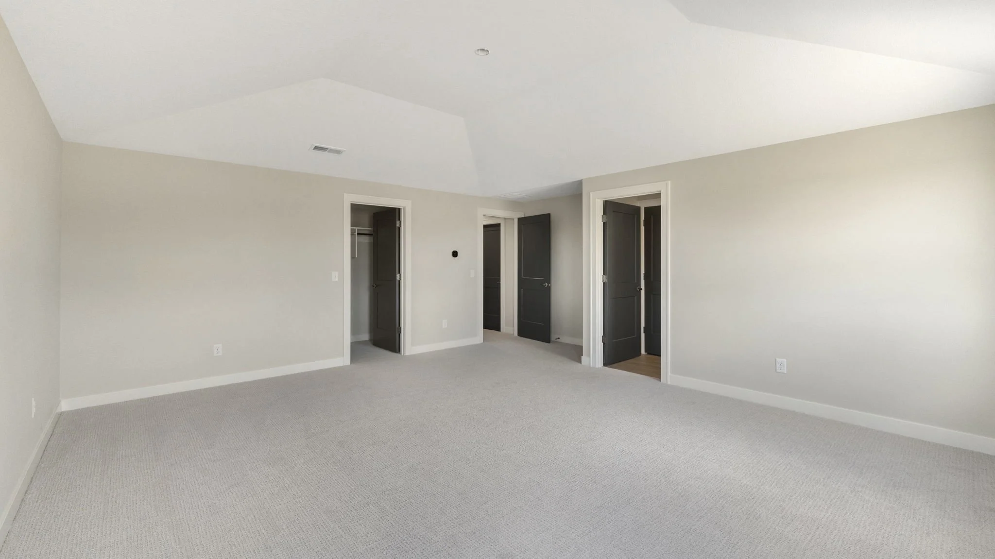 Empty room with light-colored walls, beige carpet, and three open closet doors, two painted dark gray and one white.