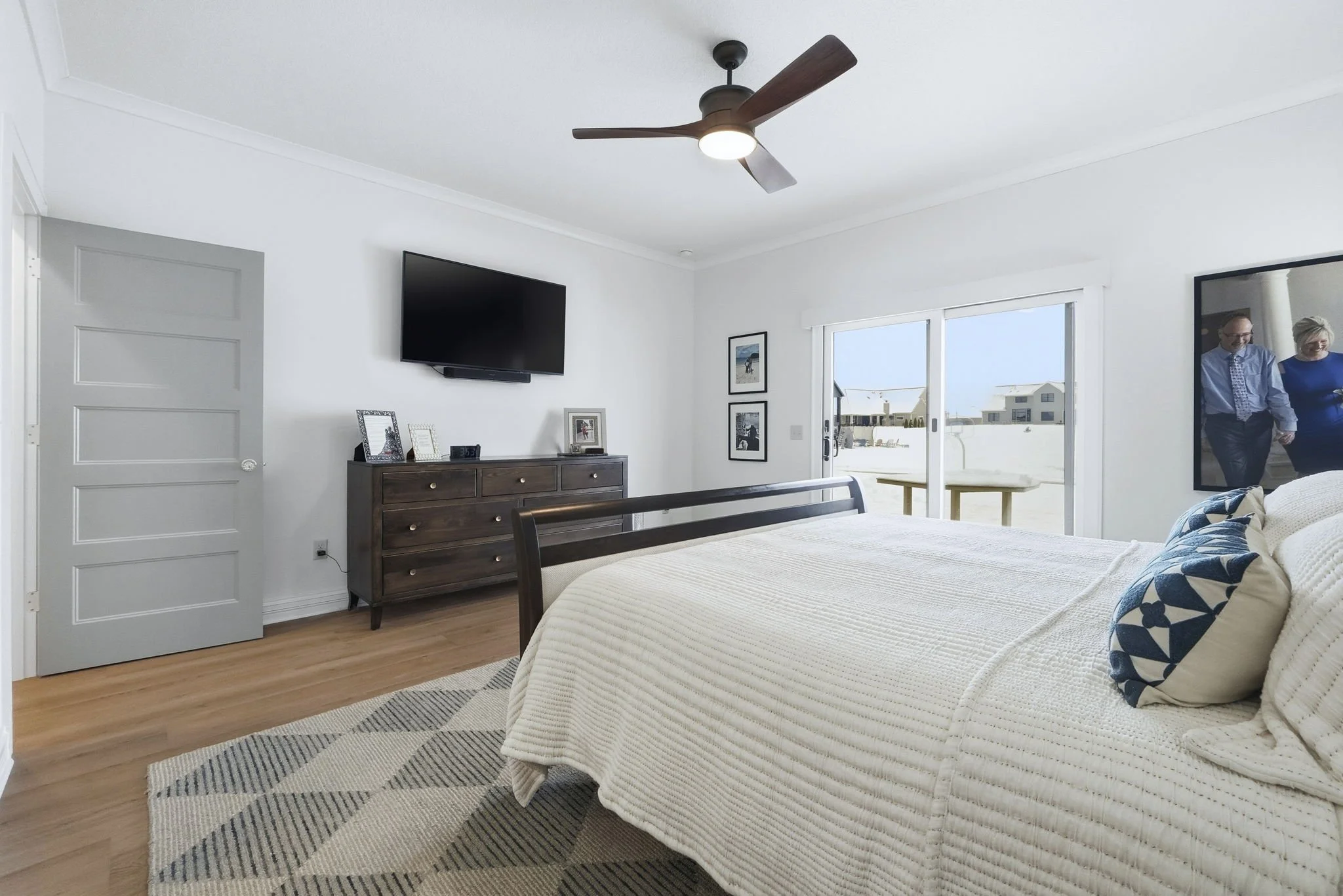 A bedroom with a large bed covered in a cream textured blanket and decorative pillows, a dark wooden dresser with picture frames and decor, a wall-mounted flat-screen TV, a ceiling fan with wooden blades, chairs outside visible through sliding glass 