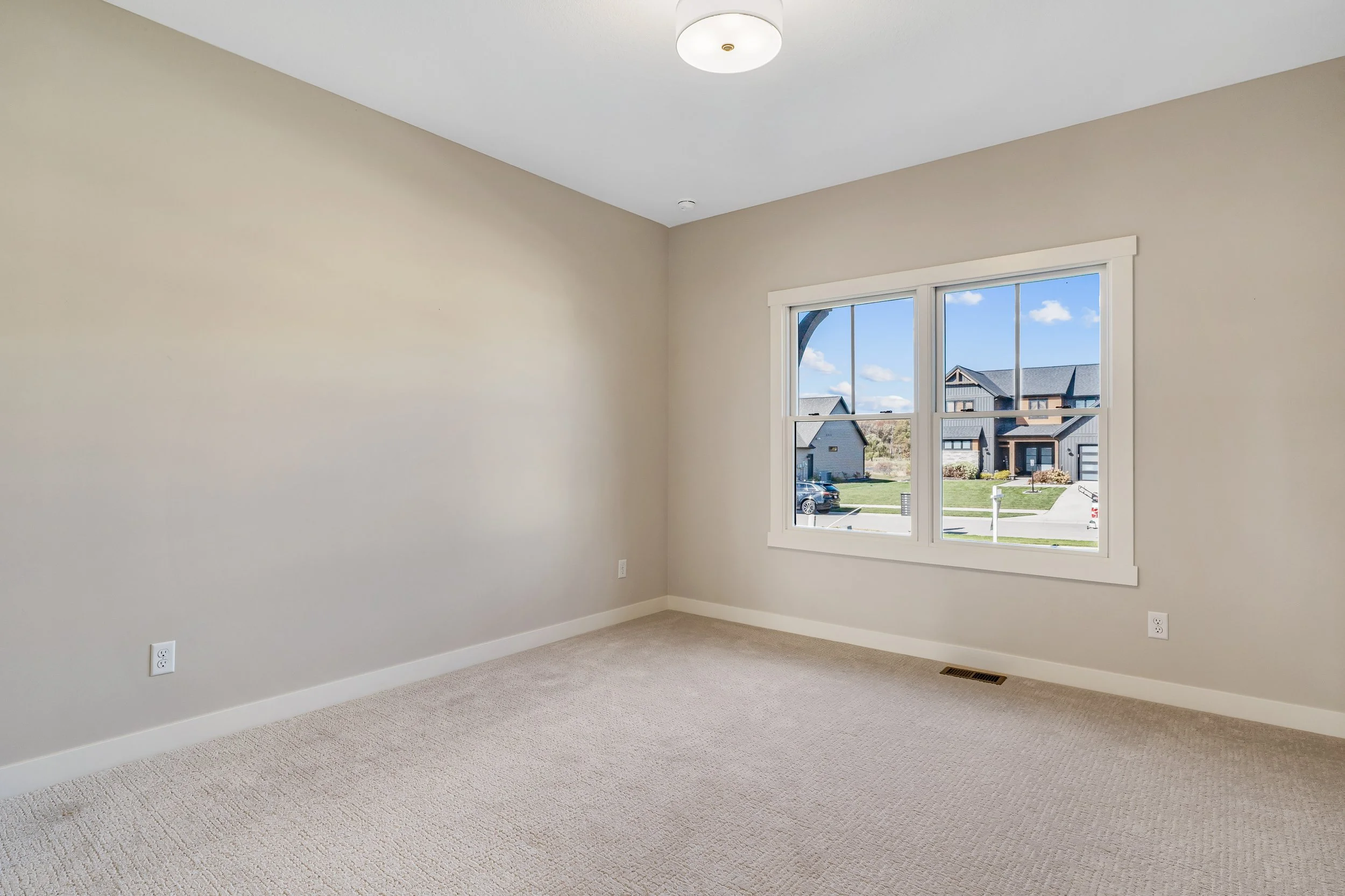 Empty room with beige carpet and light-colored walls, large window showing houses and sky outside, ceiling light fixture.