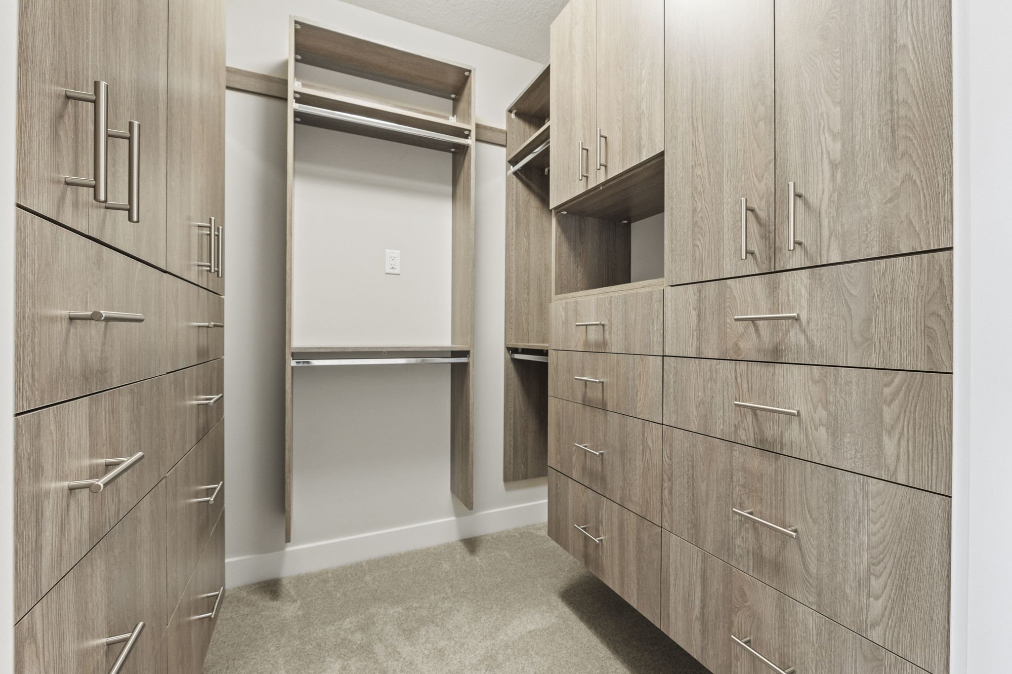 Empty walk-in closet with light wood cabinets, drawers, and open shelving, beige carpet, and a wall outlet.
