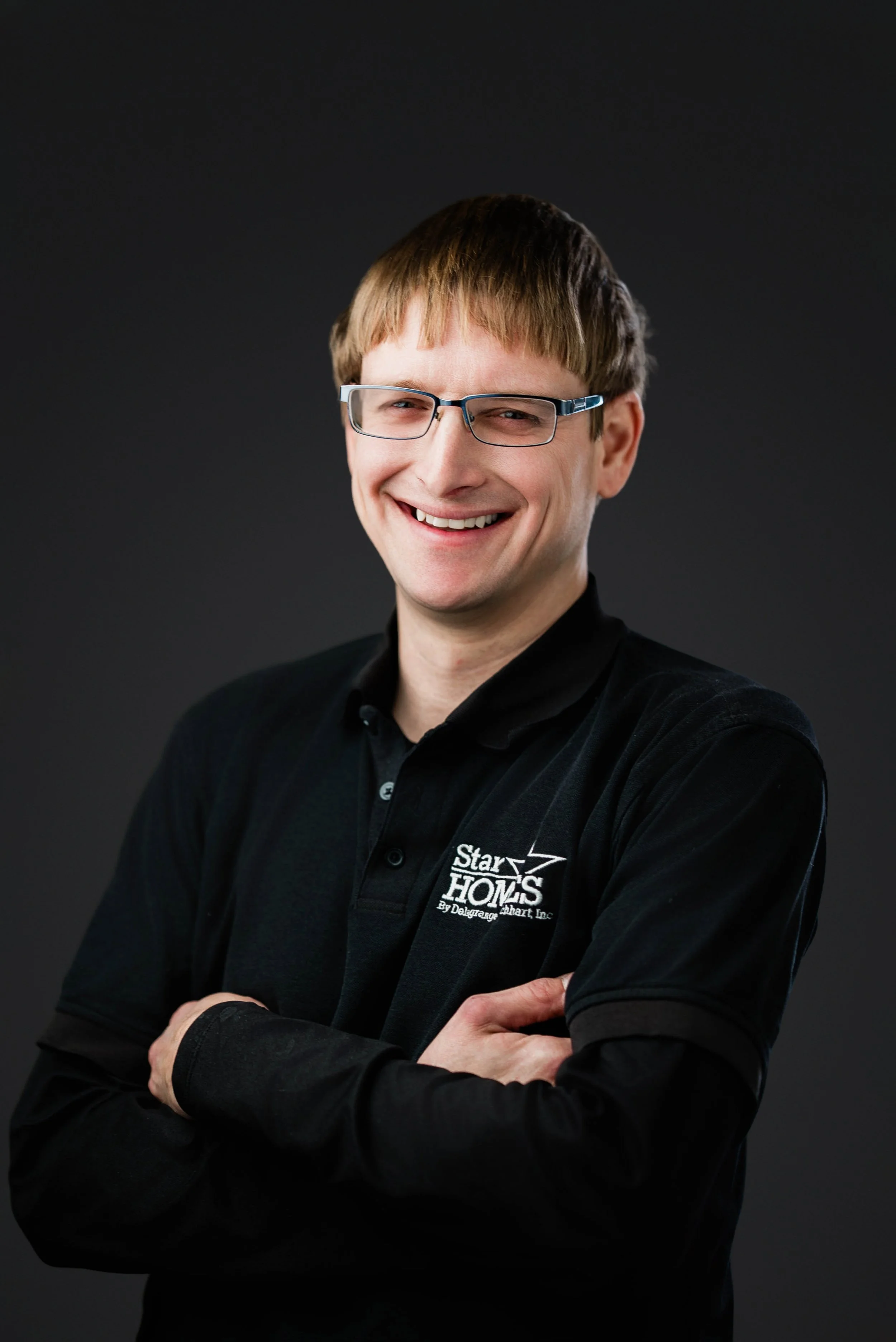 Man with light brown hair, wearing glasses and a black polo shirt with a logo, smiling with arms crossed against a dark background.