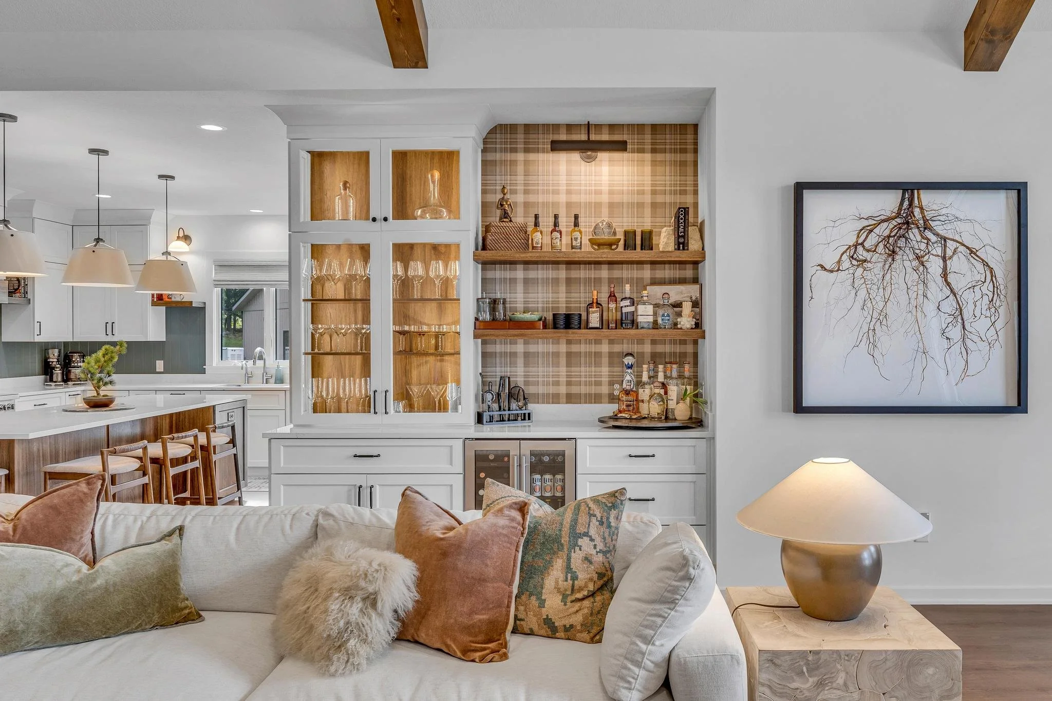 Living room with white sofa, decorative pillows, wooden side table with lamp, white cabinetry with glassware, open bar area with shelves of liquor bottles, and a framed artwork of tree roots on the wall.