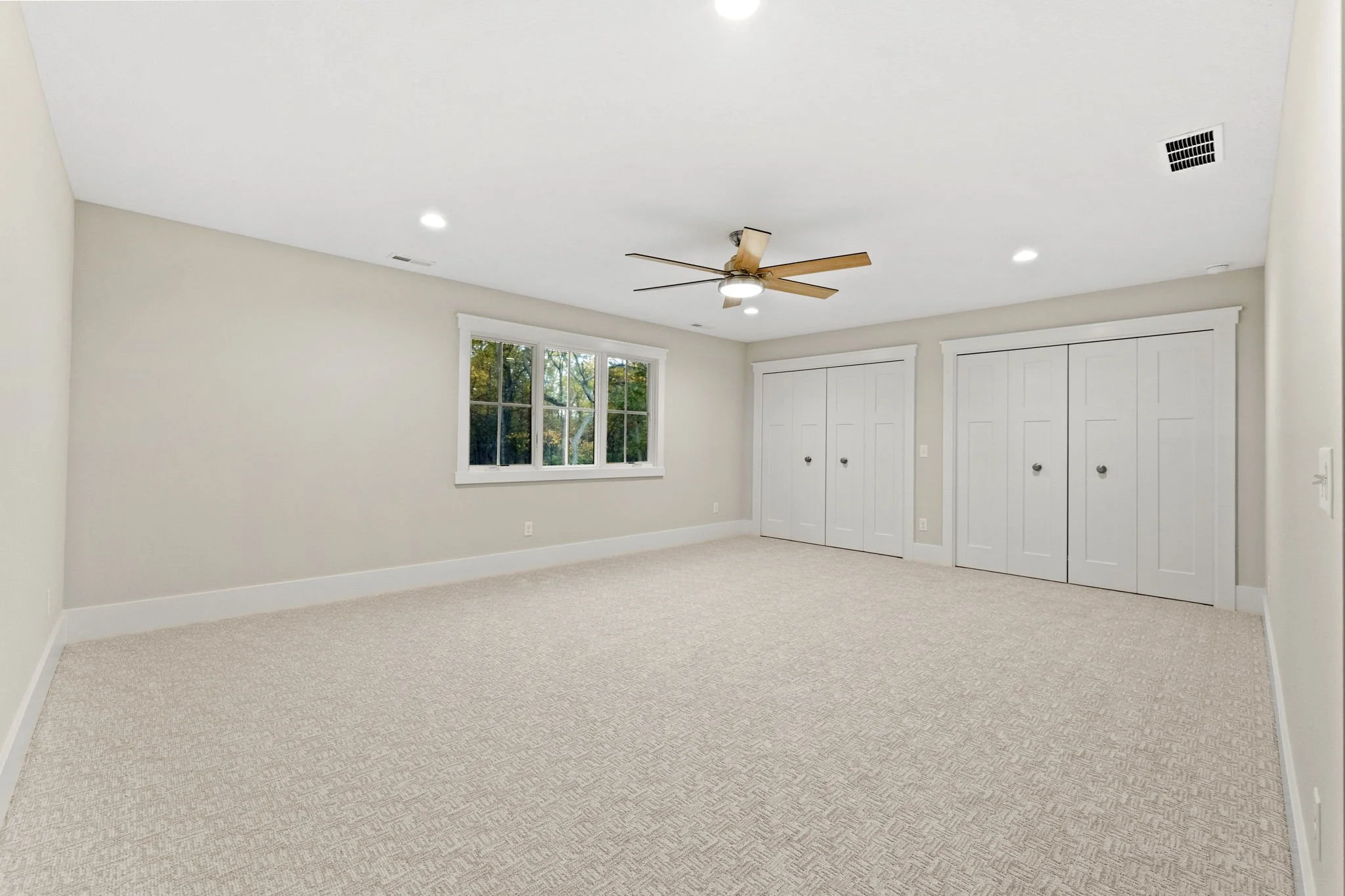 Empty bedroom with beige carpet, two white closet doors, a window showing trees outside, a ceiling fan with wooden blades, and recessed lighting.