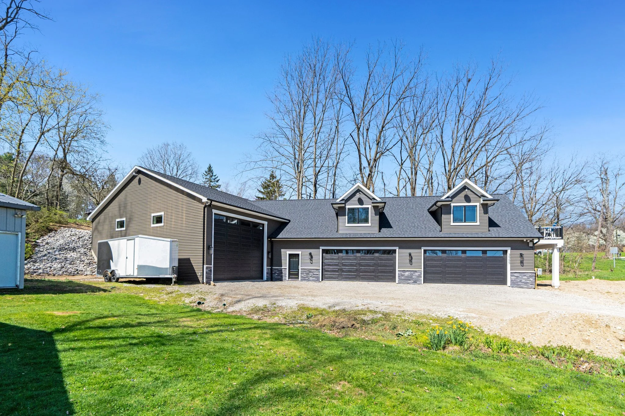A modern house with a large garage, surrounded by a green lawn and leafless trees in the background on a clear, sunny day.