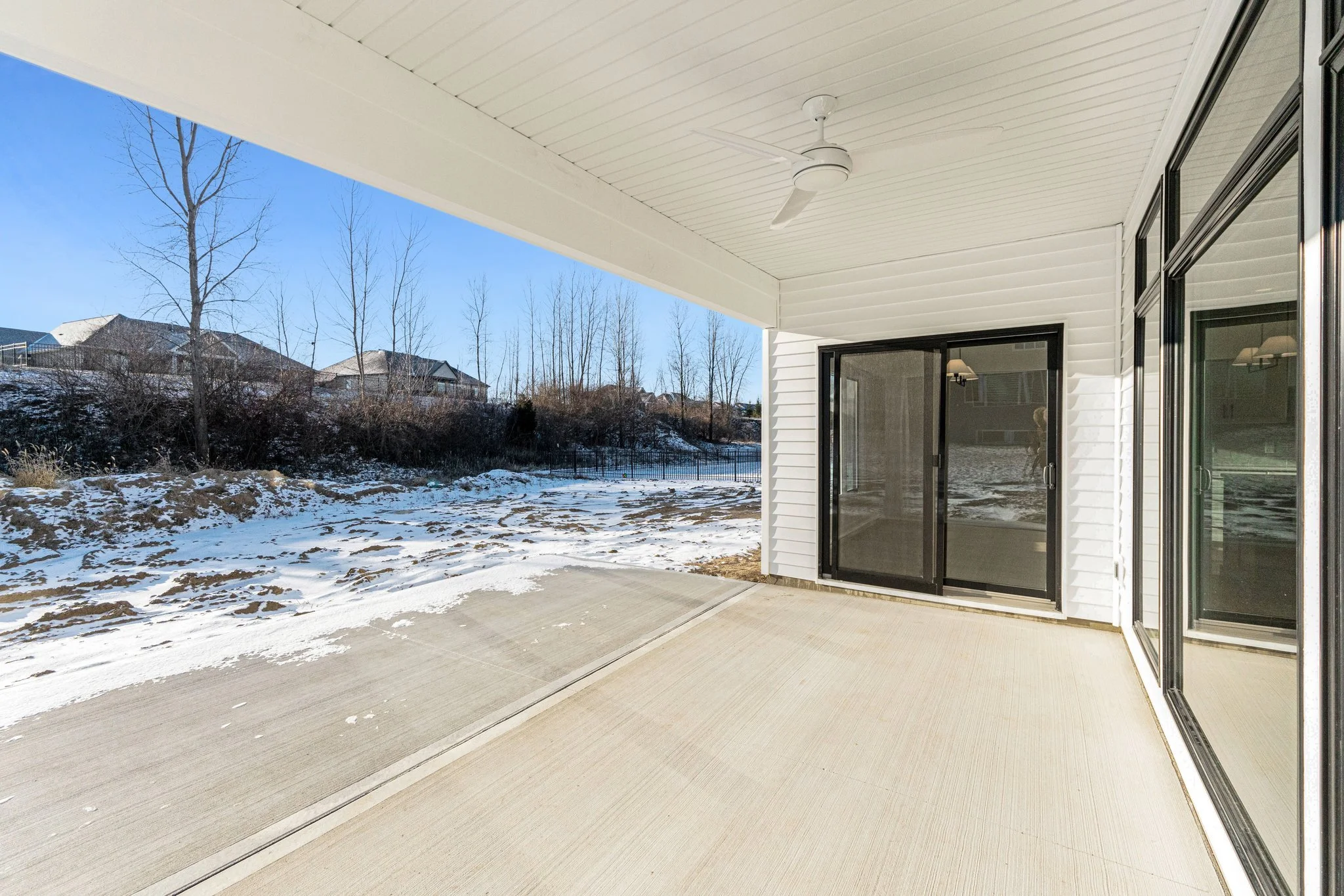 Empty covered patio with white ceiling and walls, ceiling fan, sliding glass doors, and a view of a snowy backyard with trees and houses in the background.