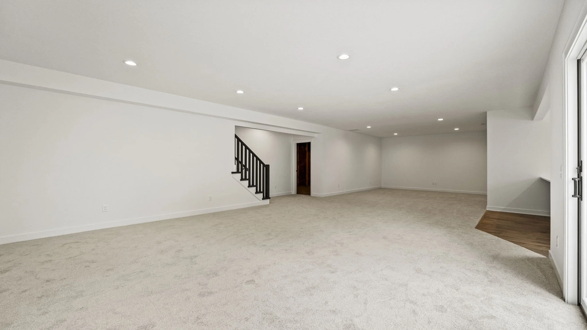Empty living room with white walls, beige carpet, ceiling lights, and a staircase with black railing.