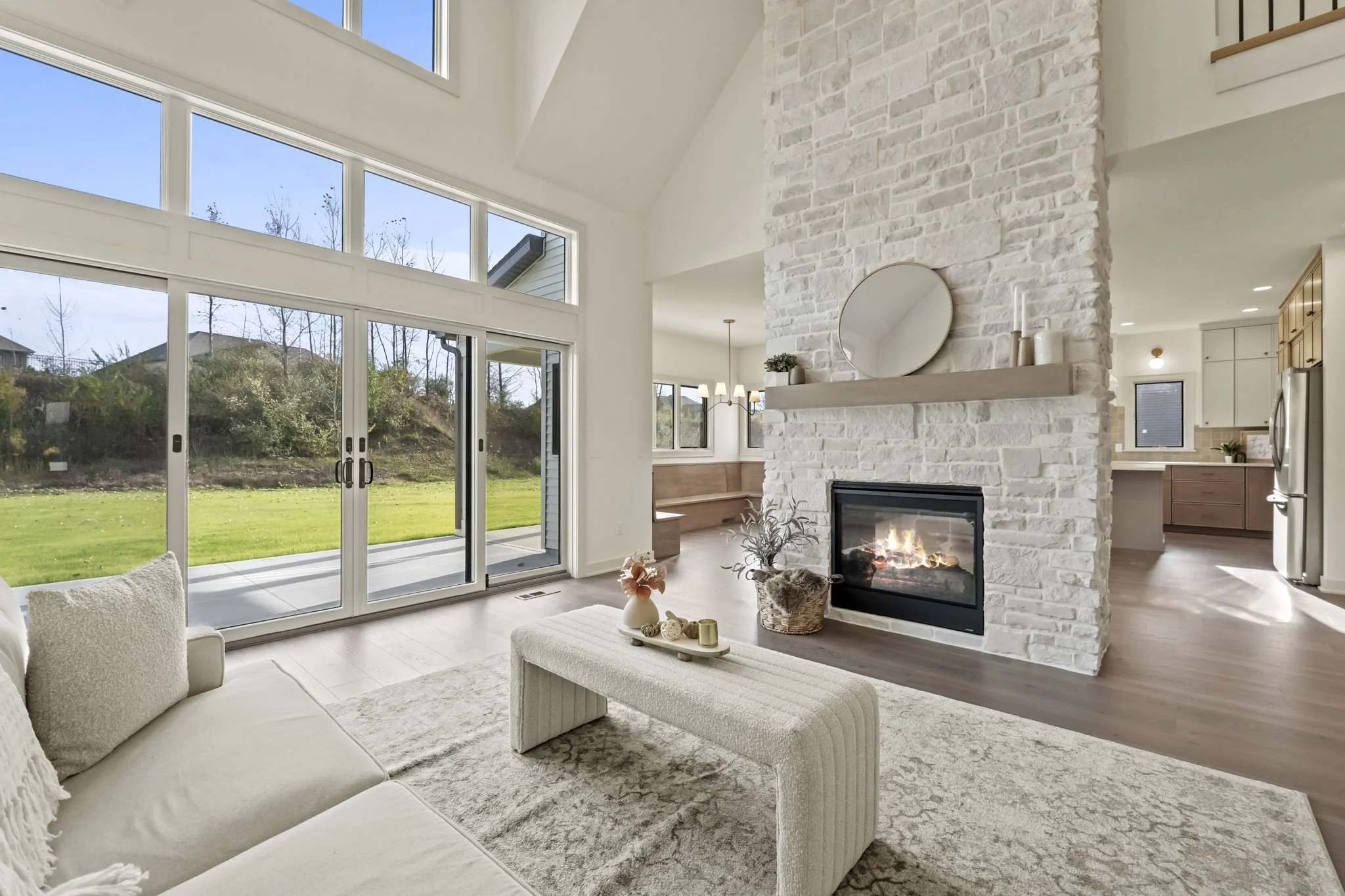Living room with white sofa, beige rug, and light-colored coffee table. Large sliding glass door opens to a grassy backyard. White brick fireplace with round mirror and decor on mantel. Bright, airy space with high vaulted ceiling and large windows.
