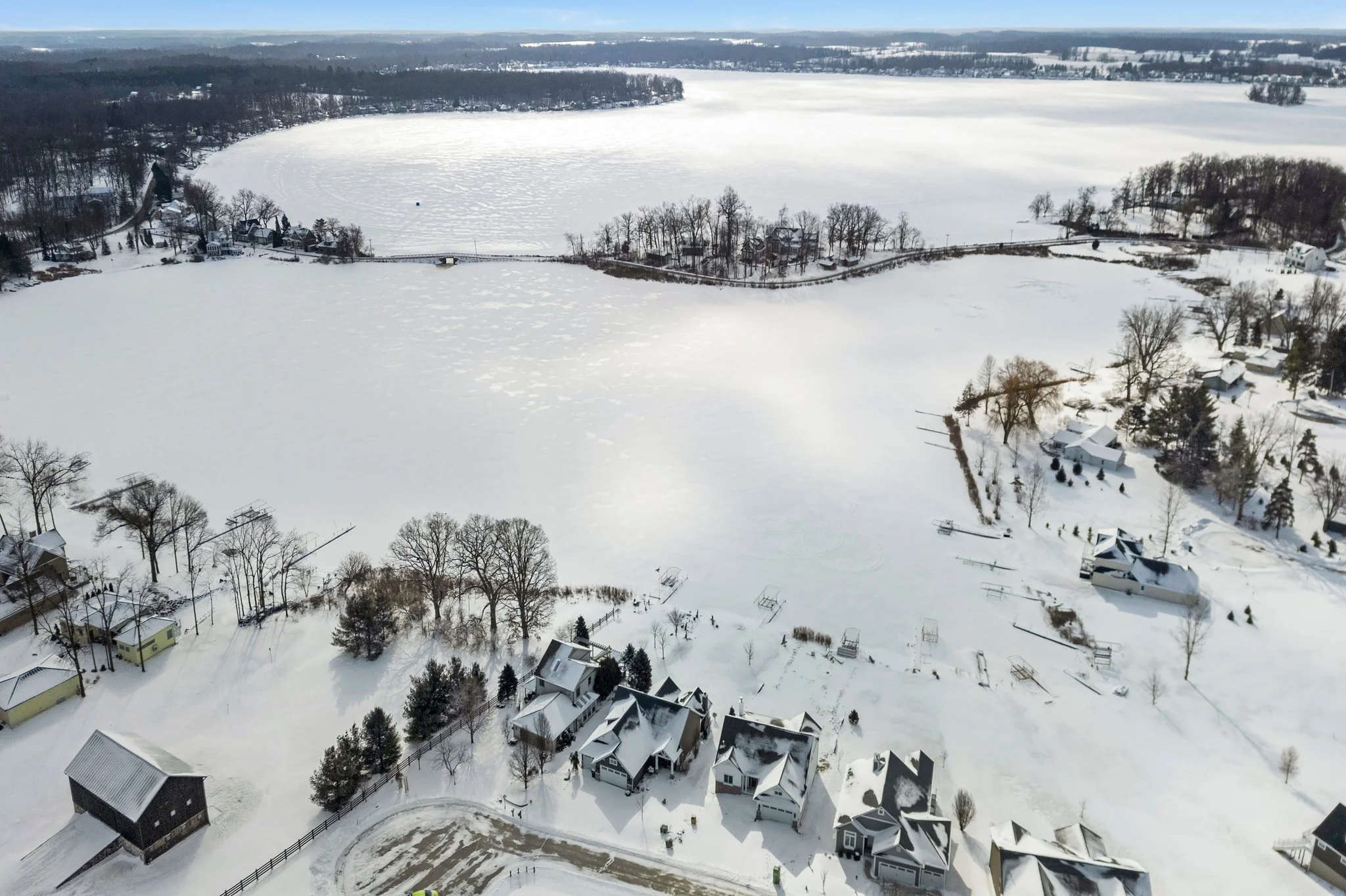 A snow-covered lake with houses and trees on the shoreline, seen from above during winter.