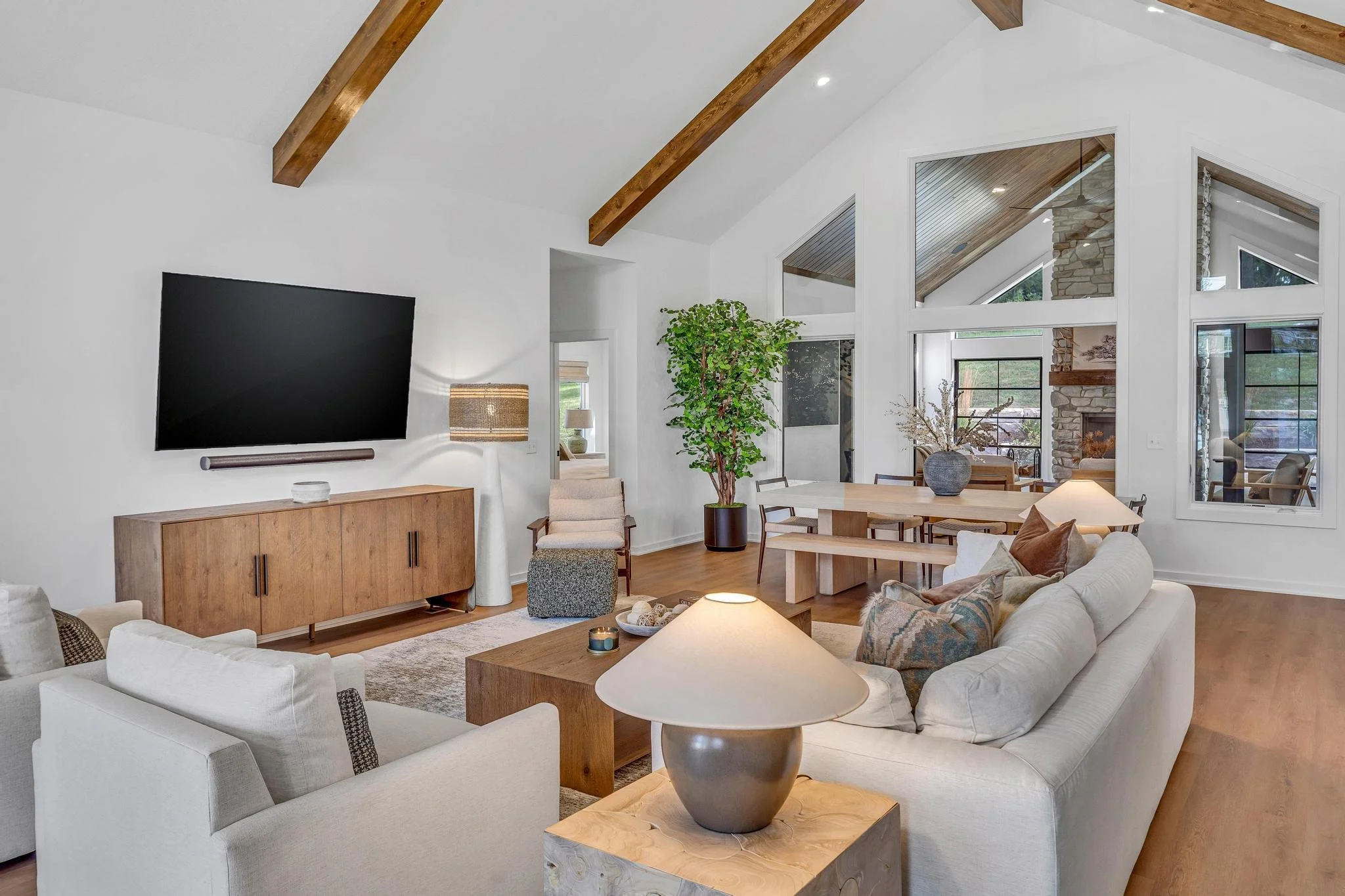 Living room with white walls, vaulted ceiling with exposed wooden beams, large windows, a flat-screen TV on a wooden console, beige sofas, a wooden coffee table, a side table with a lamp, a potted plant, and a dining area in the background.