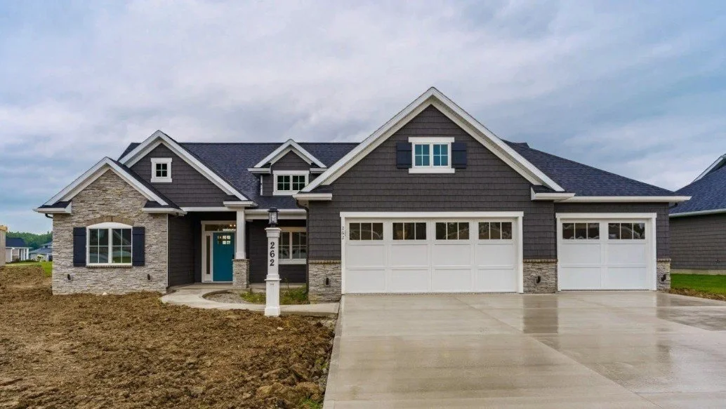 Newly built house with gray siding, stone accents, a three-car garage, and a concrete driveway, located in a suburban neighborhood.