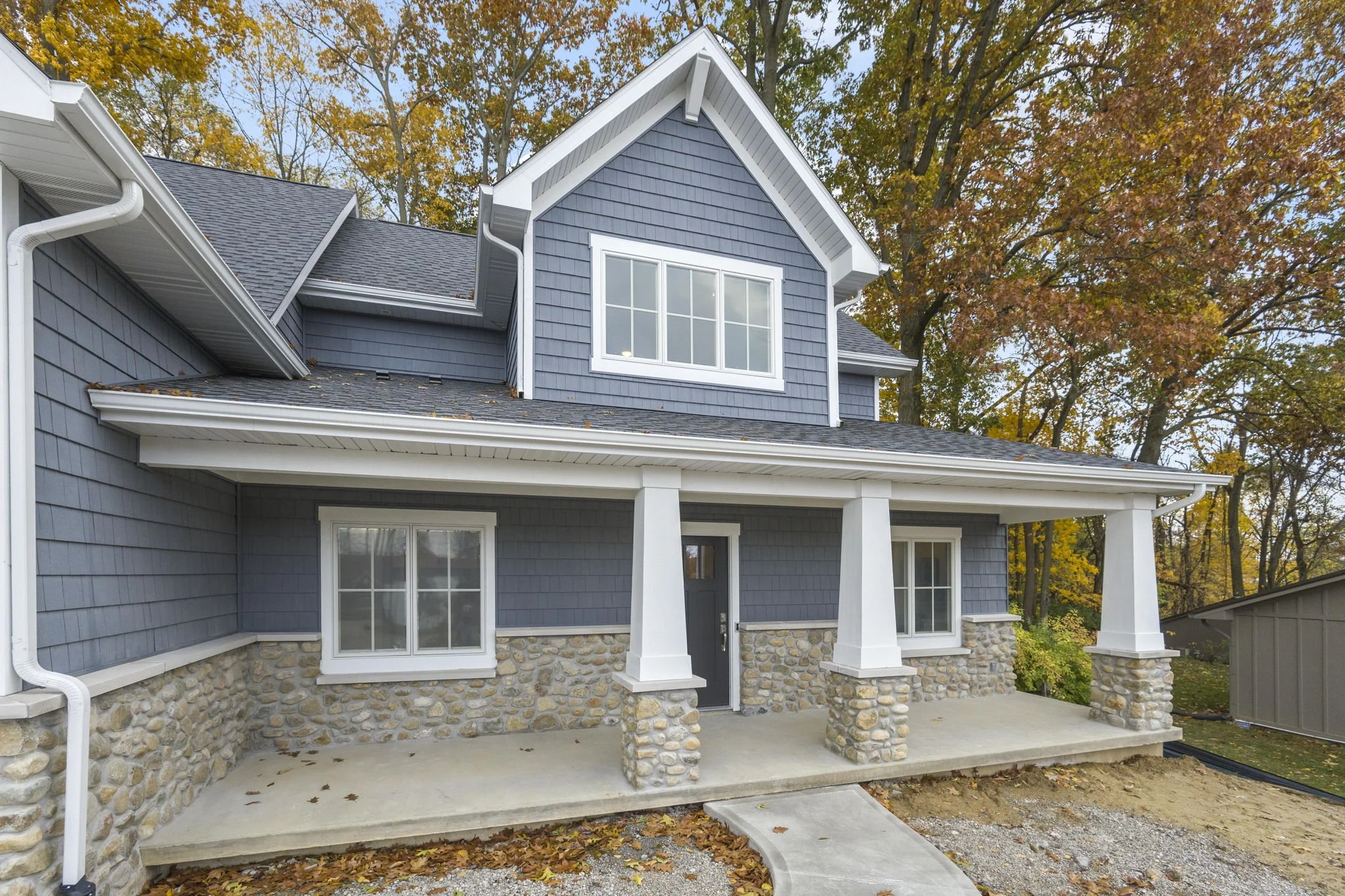 Front view of a two-story house with dark blue siding, stone foundation, white trim, and a covered porch with four white columns, surrounded by autumn trees.