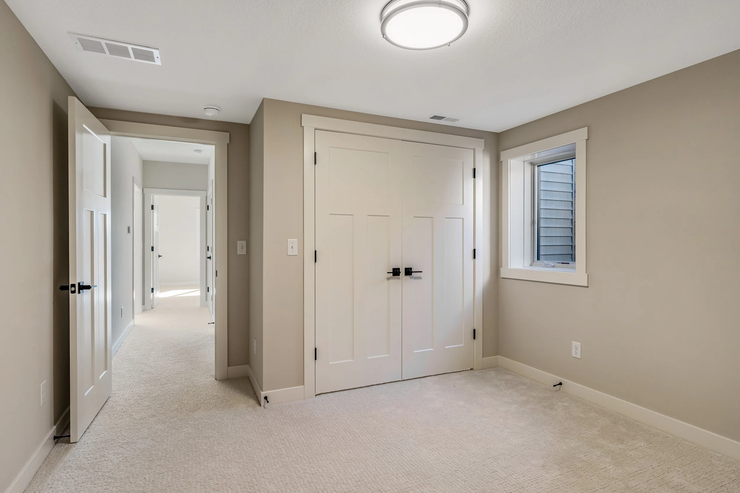 Empty bedroom with beige walls, white trim, a window, ceiling light, door open to hallway, closet with double doors
