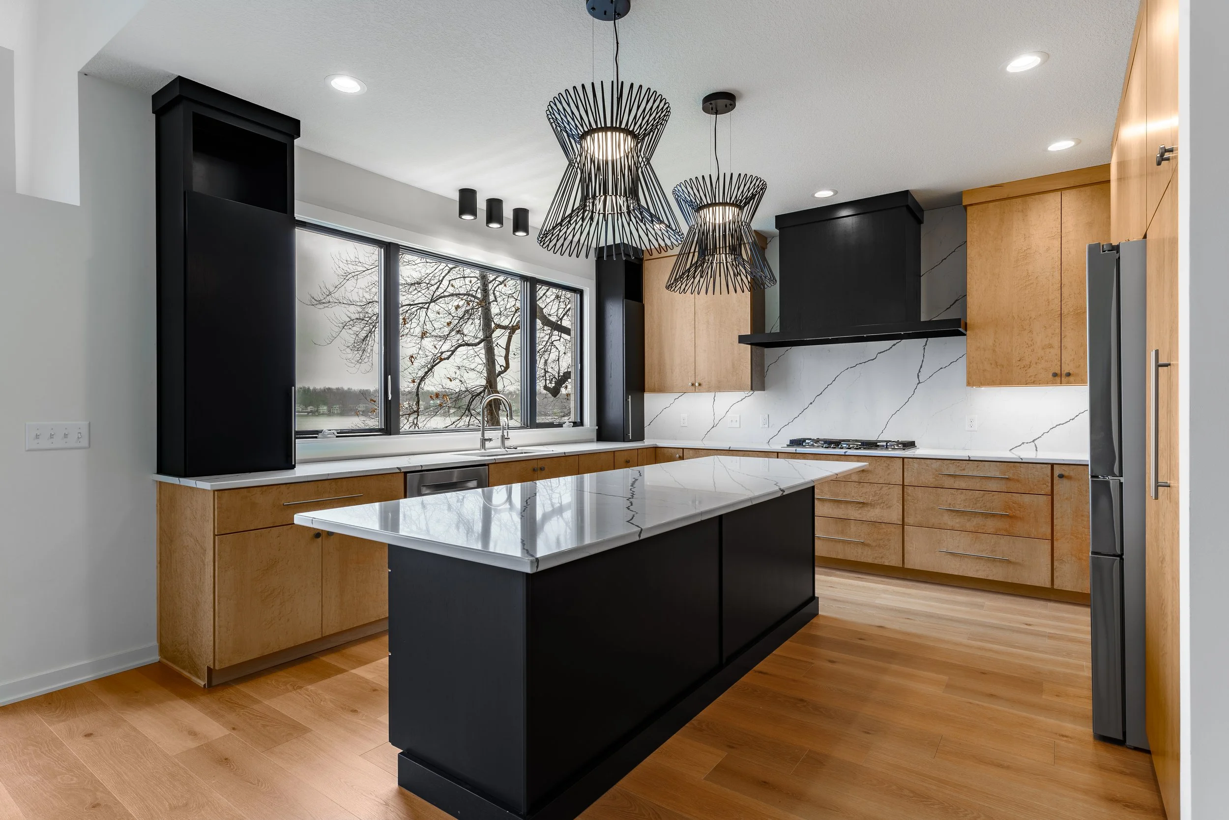 Modern kitchen with black and natural wood cabinets, a large marble island, a stainless steel refrigerator, black geometric pendant lights, and a large window showing an outdoor view.
