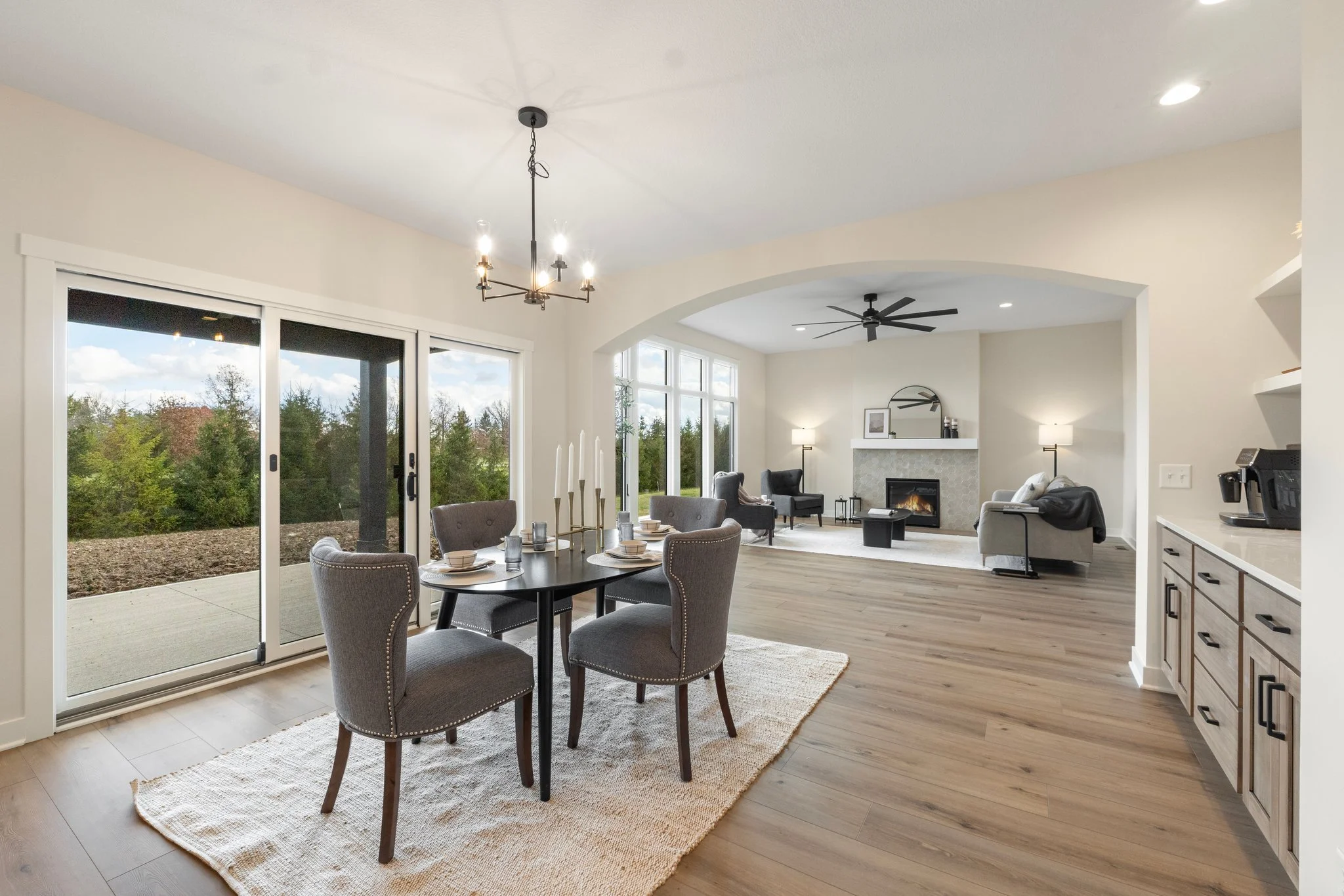 Open-concept living and dining room with large sliding glass doors, light-colored walls, hardwood floors, a black dining table with four gray upholstered chairs, a cozy sitting area with a fireplace, and modern ceiling fans and lighting.