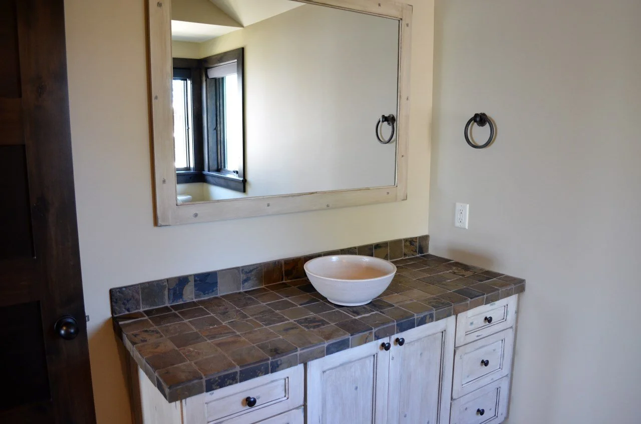 Bathroom vanity with a tiled countertop, a white vessel sink, a large mirror with a wooden frame, and two towel rings on the wall.