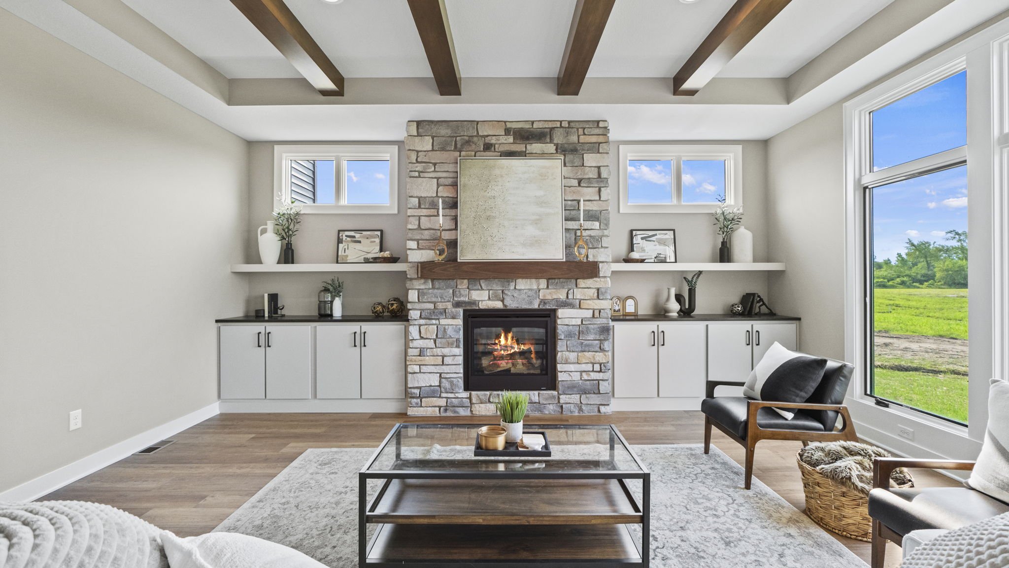 Living room with a stone fireplace, white cabinetry, and large windows showing a green outdoor landscape.