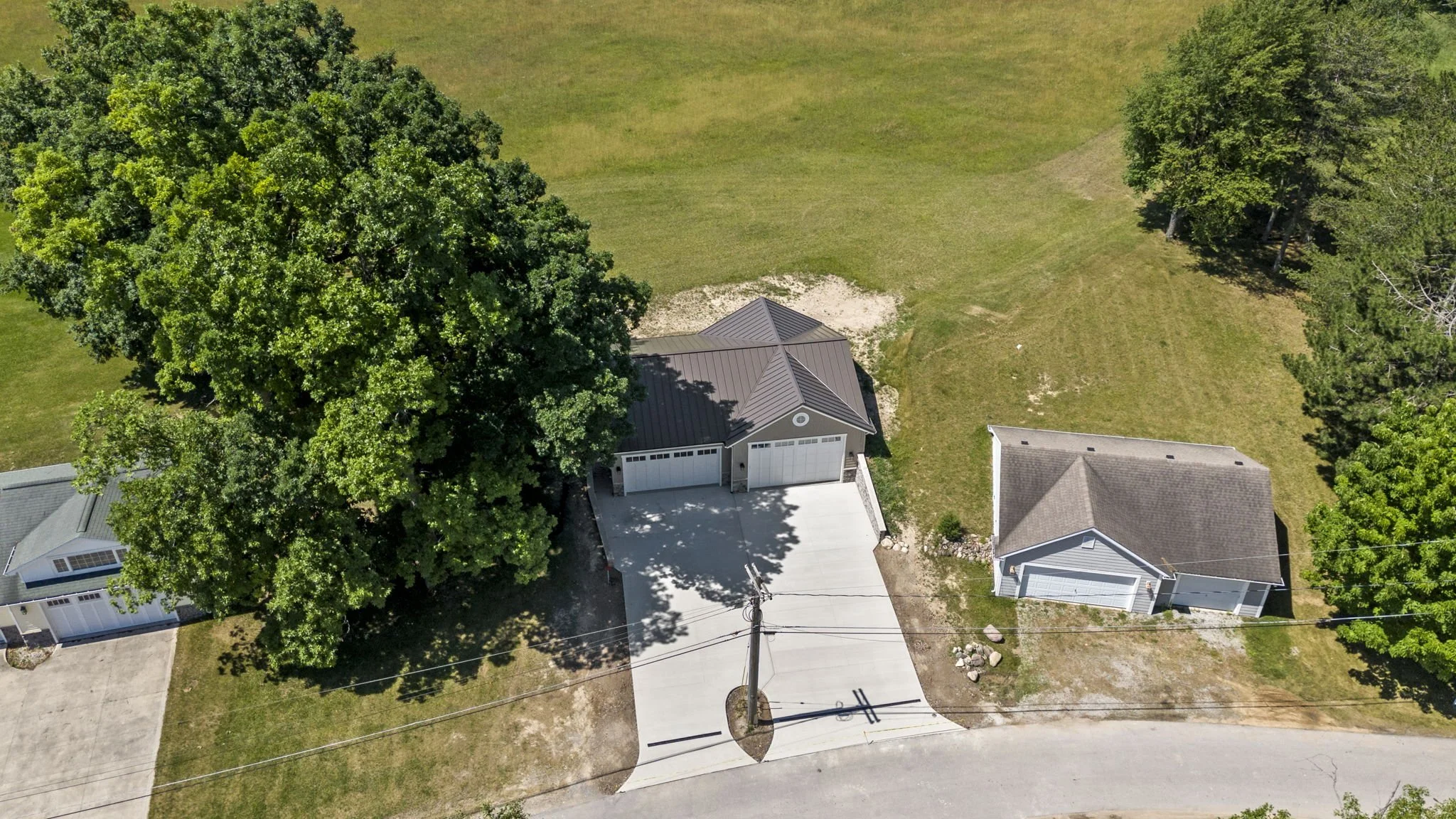 Aerial view of a residential property showing a driveway, two garage buildings, large trees, grass, and neighboring homes.
