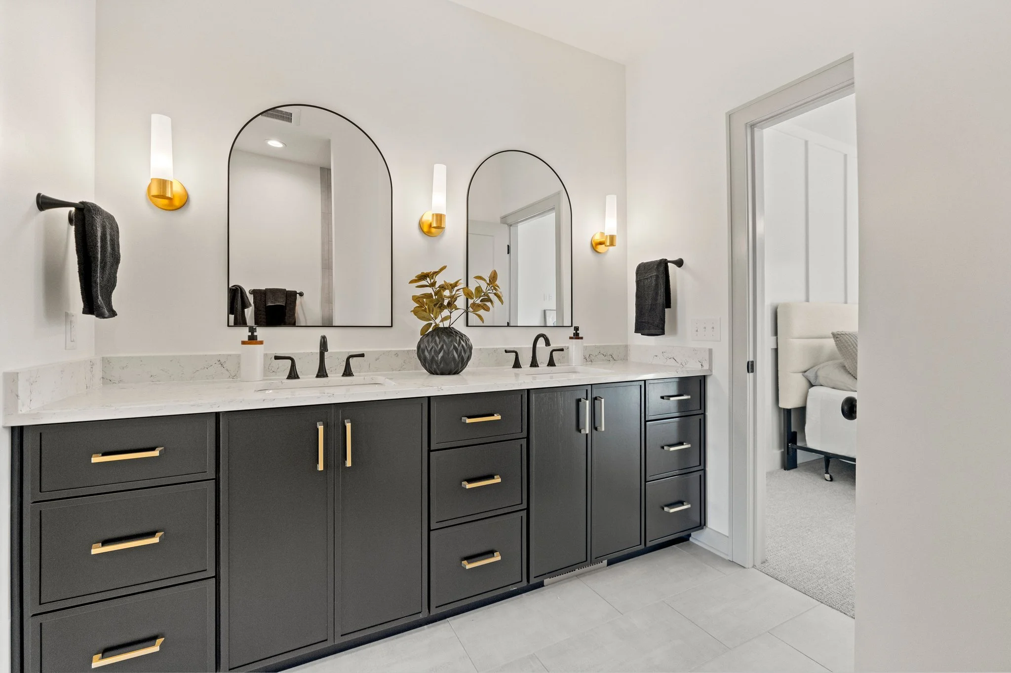 Modern bathroom with double vanity, black cabinets with gold handles, white marble countertop, two oval mirrors, wall-mounted light fixtures, black towels, and a doorway leading to a bedroom.