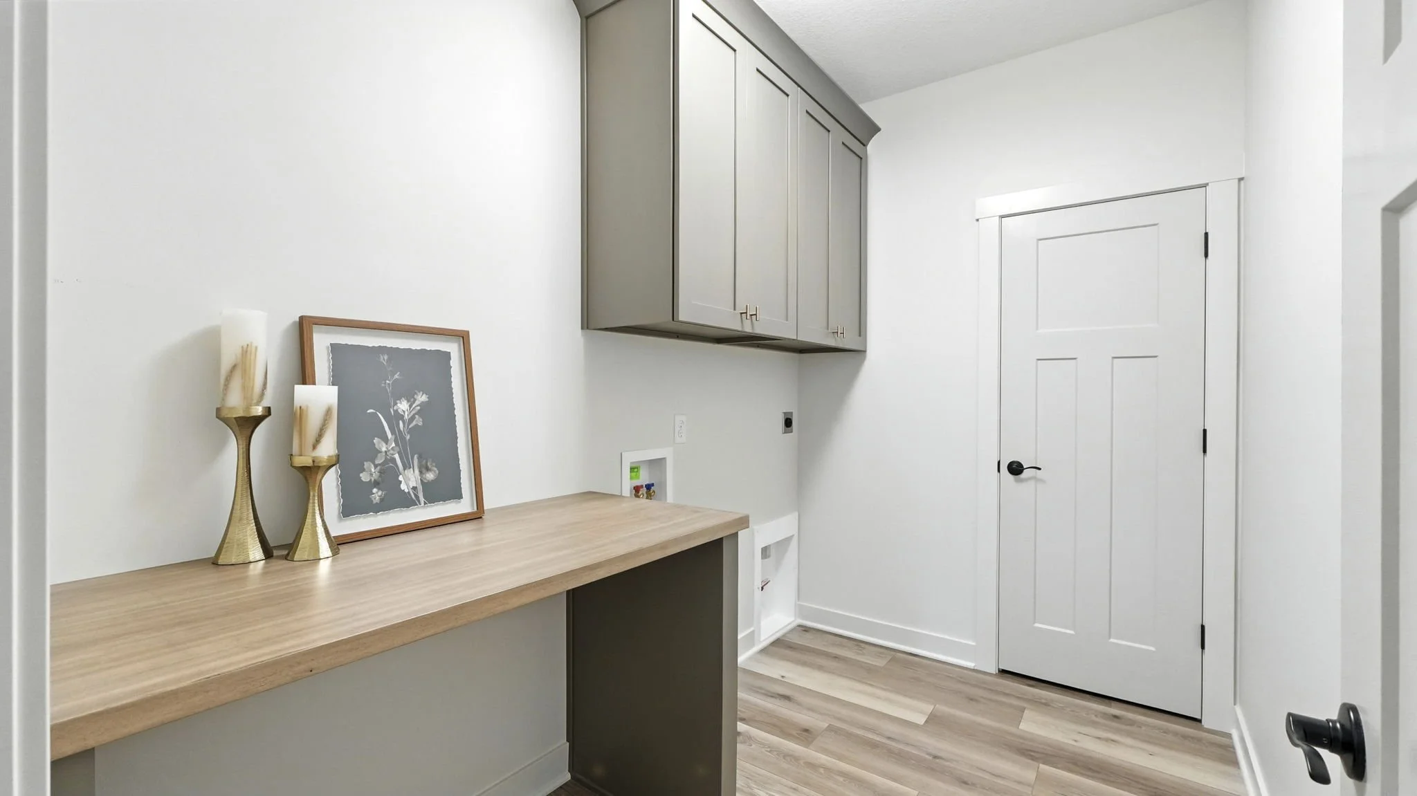 Interior view of a laundry room with gray cabinets, a wooden countertop, decorative candles, and artwork. White walls and a closed door are visible.