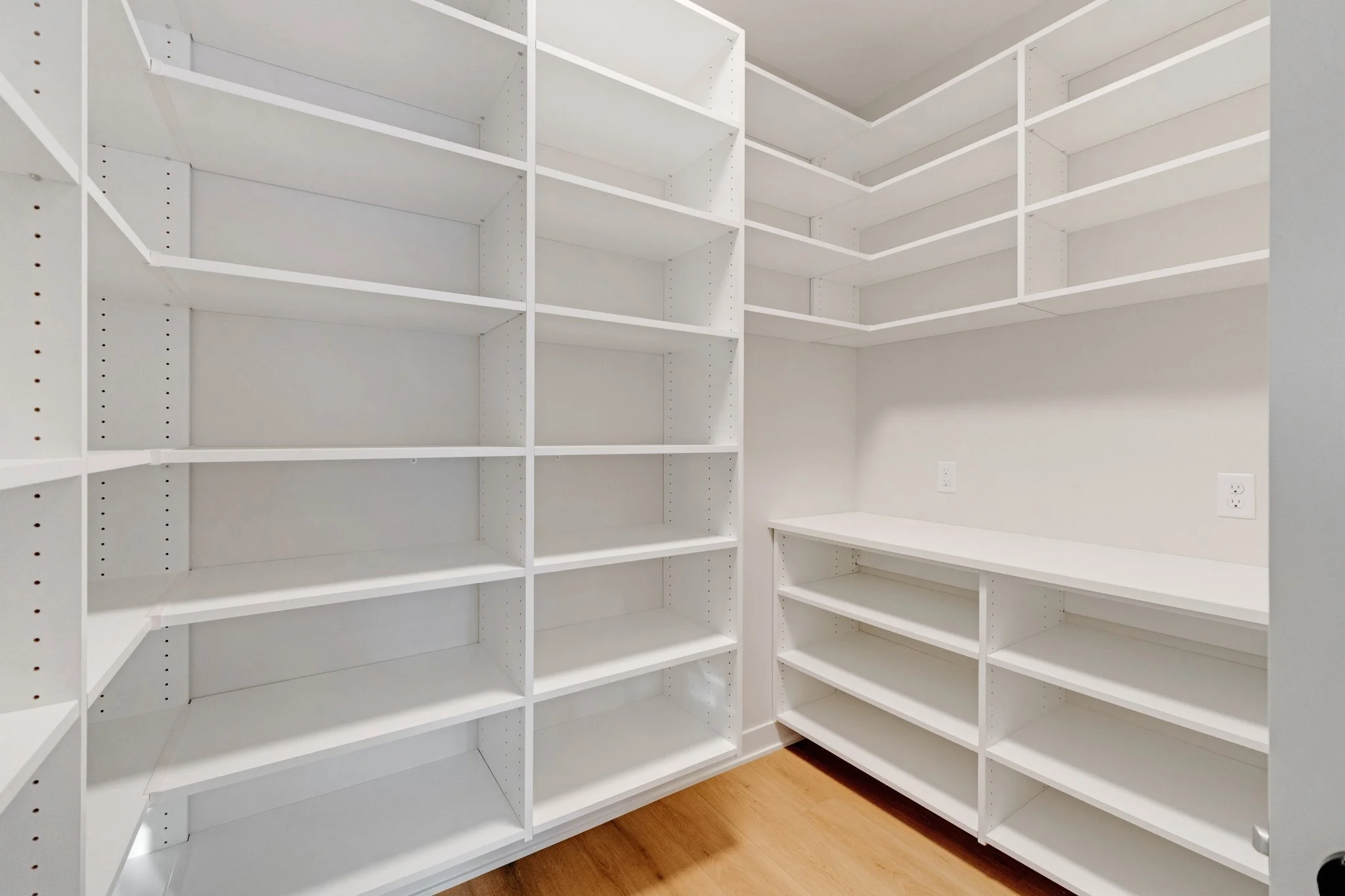 Empty white built-in shelves and cabinets in a walk-in closet with hardwood flooring.