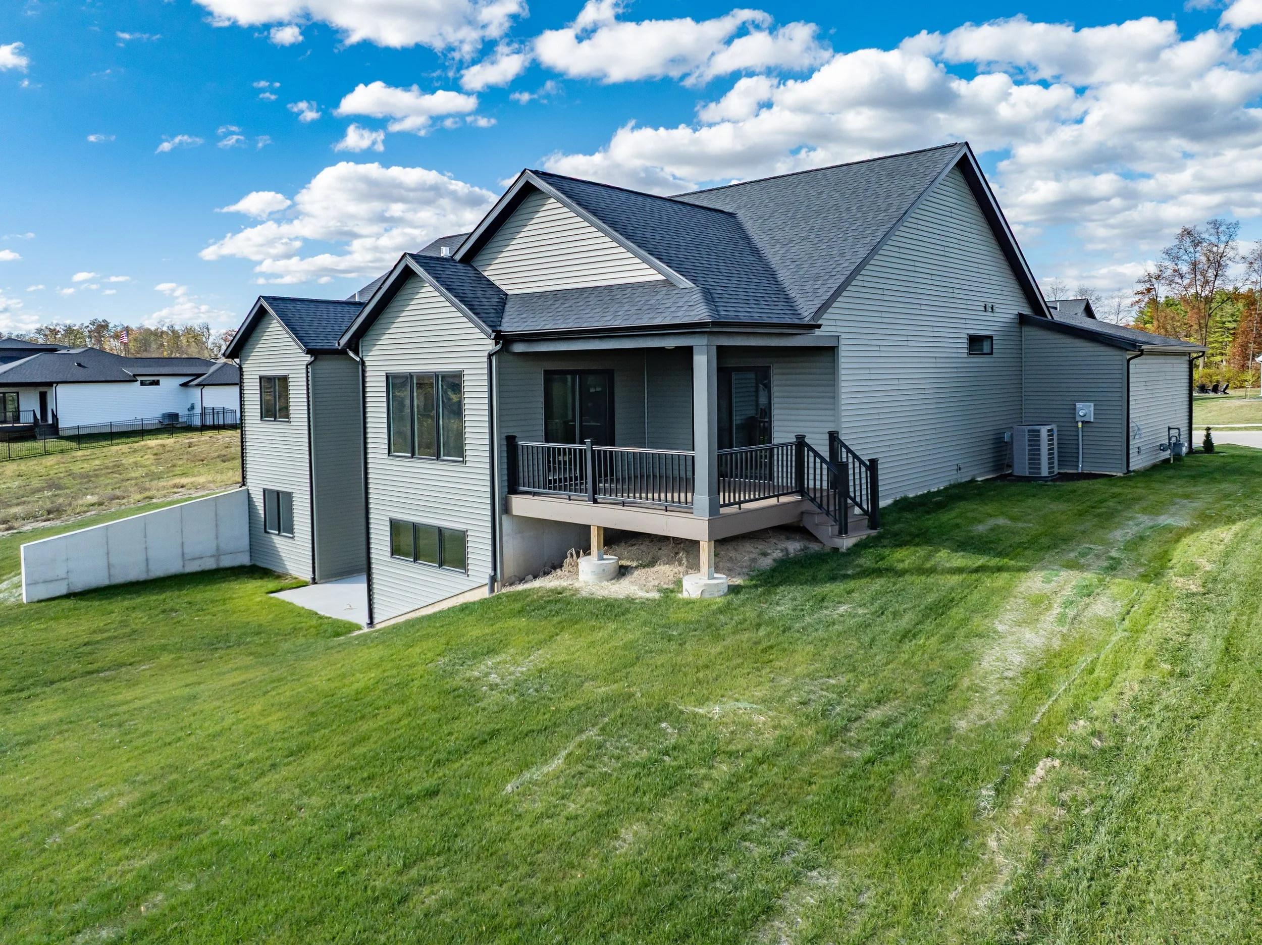 Rear view of a modern, two-story house with beige siding and a black roof, featuring a deck with black railing, on a grassy hill with neighboring houses and a partly cloudy sky in the background.