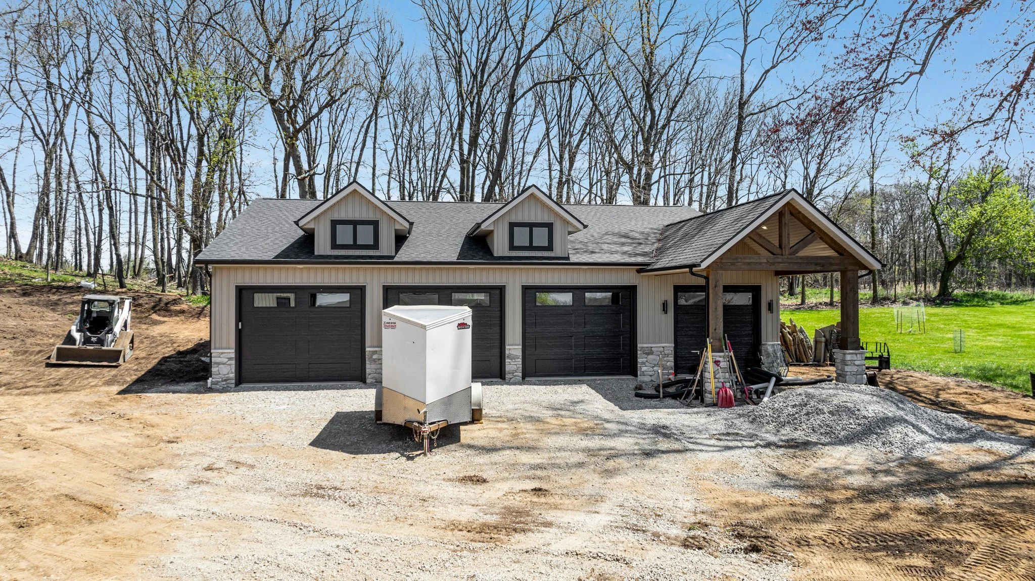 Newly built house with a three-car garage, front porch, surrounded by a wooded area and on a construction site with gravel driveway and construction equipment.