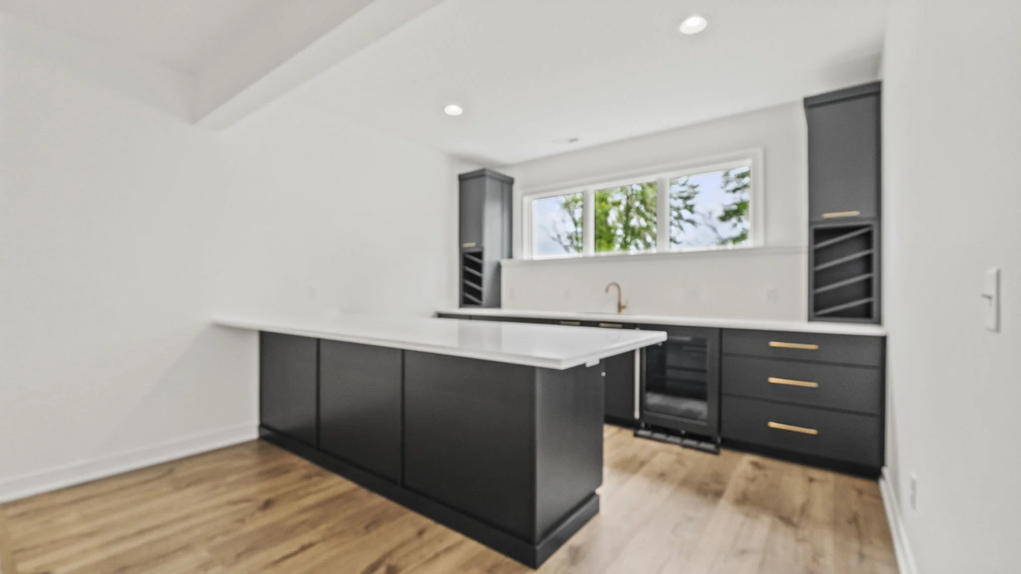 Modern kitchen with white walls, black cabinets, and gold hardware. Large window with greenery outside, wooden floor, and an island with a white countertop.