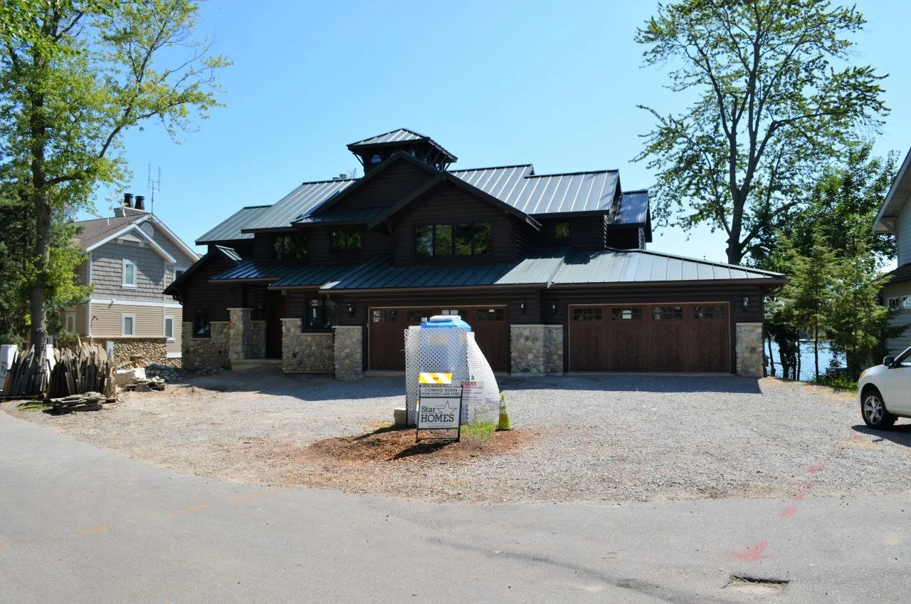 A new house under construction with a black exterior, stone accents, and a metal roof. Construction materials and a sign indicating future homes are in front of the house. There are trees and neighboring houses, with a body of water visible in the ba
