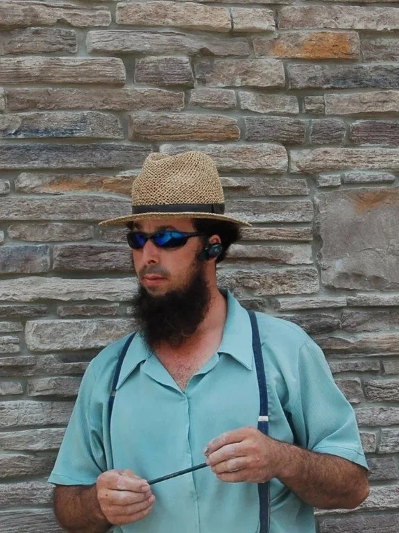 A man standing in front of a stone wall wearing a straw hat, blue sunglasses, a light blue shirt, and a headset.