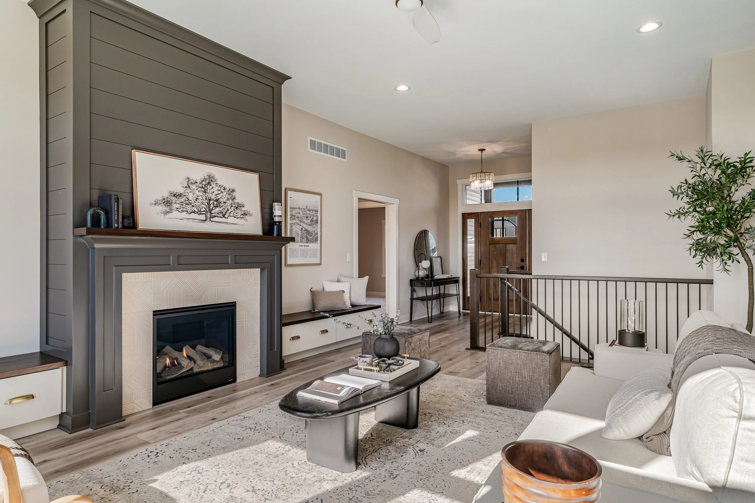 Living room with fireplace, white sofa, coffee table, decorative pillows, potted plant, and entryway with wooden door and chandelier.