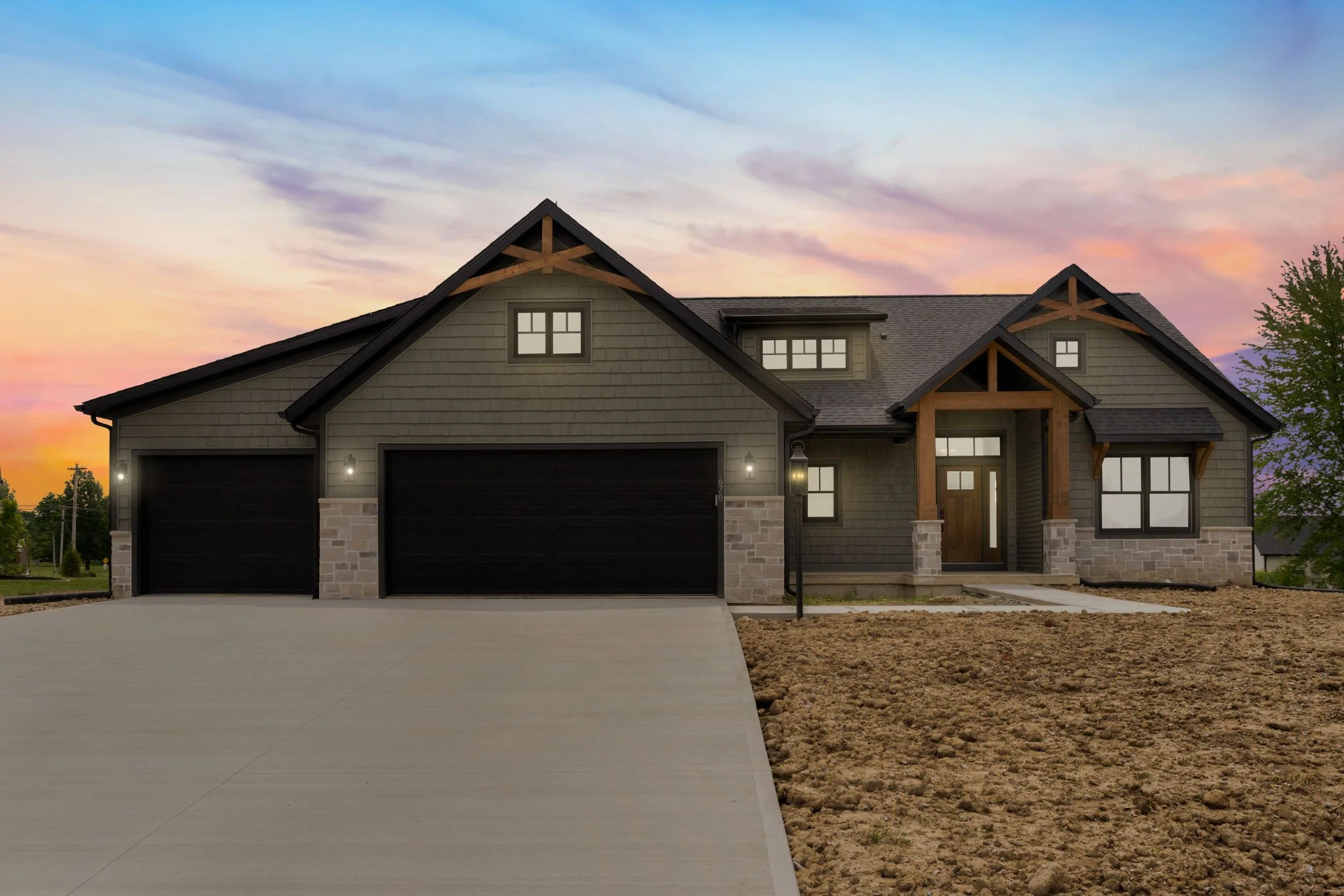 Modern house with a large driveway, stone and dark siding exterior, wooden accents, multiple windows, and a landscaped yard during sunset.