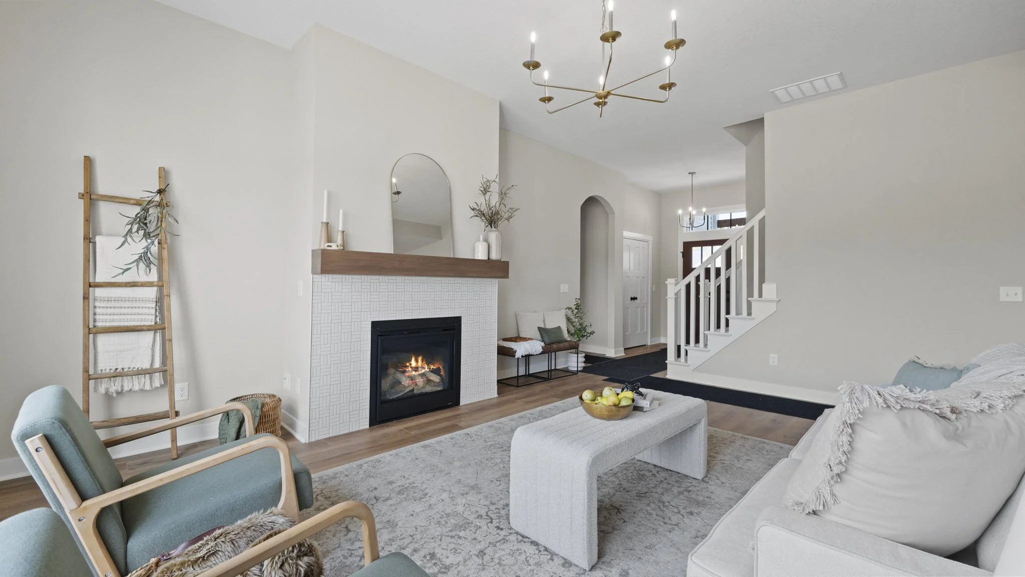 Modern living room with fireplace, white walls, wooden flooring, and contemporary furniture including chairs, a coffee table with apples, and a sofa with a blanket. There are decorative vases, a ladder with a blanket, and ceiling lights.
