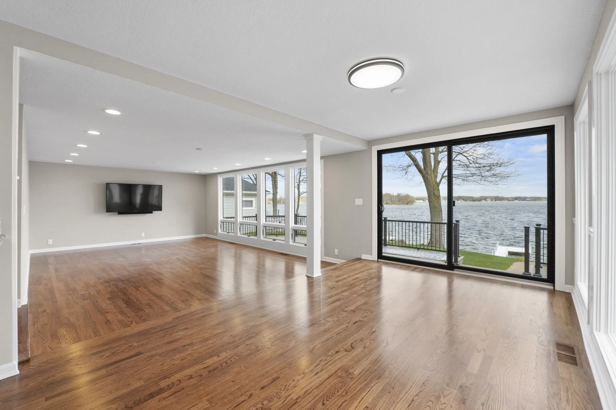Spacious living room with hardwood floors, large windows and a sliding glass door overlooking a lake with trees.