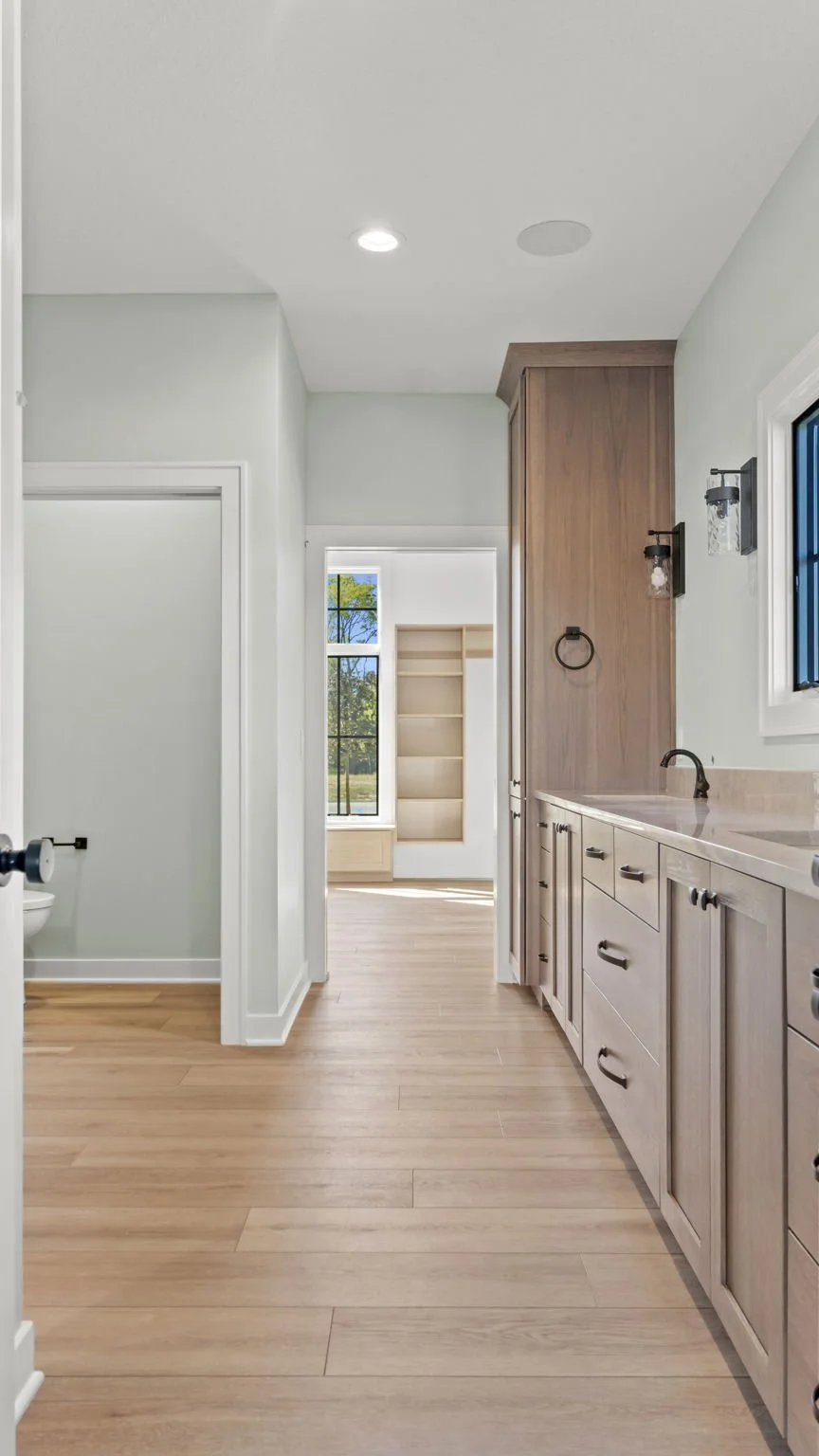 View of a kitchen with light wood cabinets, white countertops, a sink with a black faucet, and a window with a view of trees outside. Hardwood floors and a doorway leading to another room with a built-in shelf.