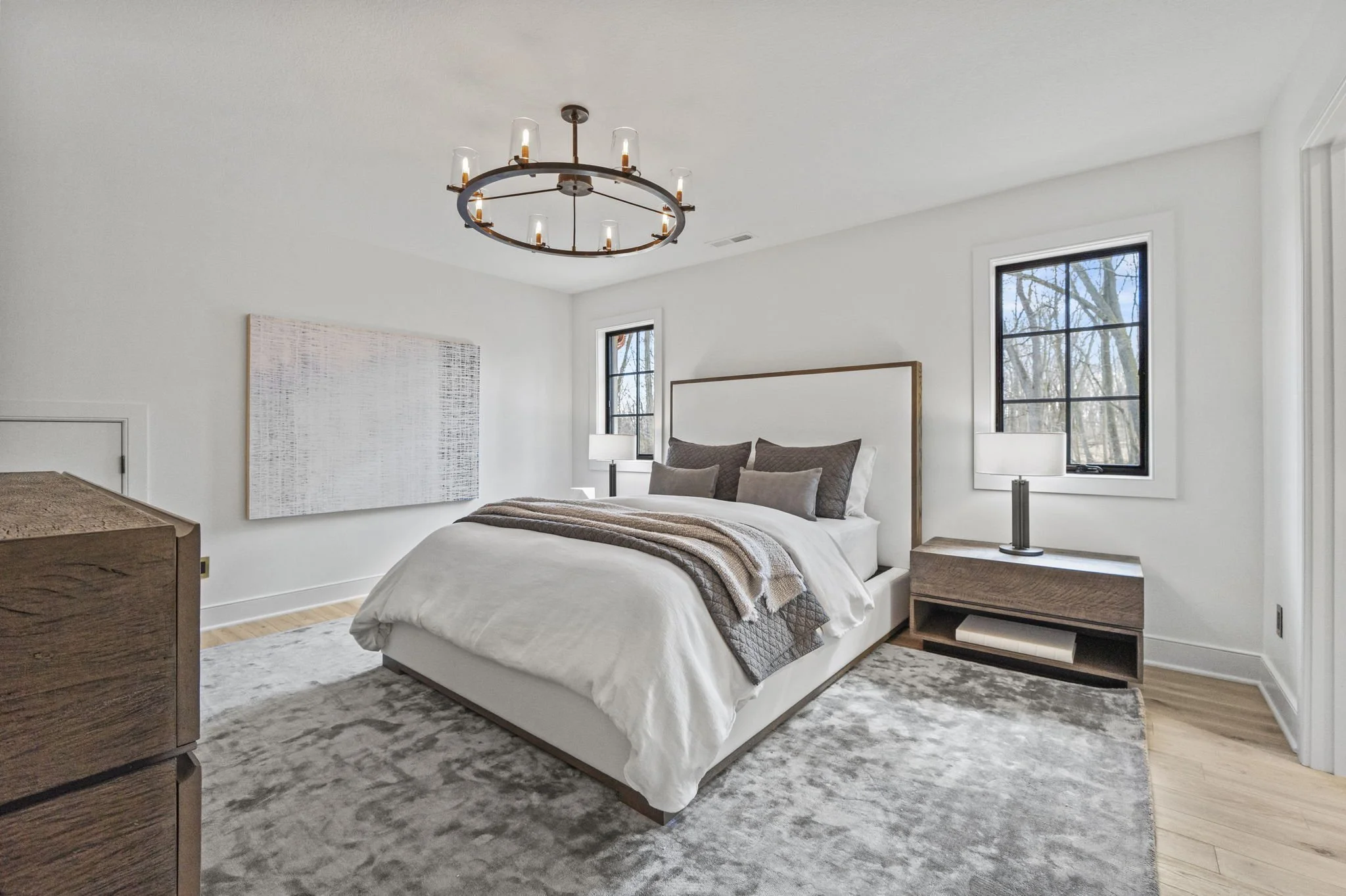 Modern bedroom with white walls, wooden furniture, and large windows showing trees outside. The bed has white linens and brown pillows, and there are lamps on either side of the bed. A chandelier hangs from the ceiling.