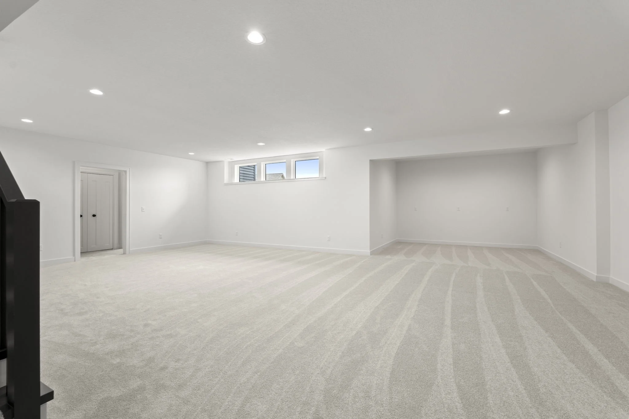 Empty, white basement room with recessed lighting and a small window, beige carpeted floor, and a closed closet door.