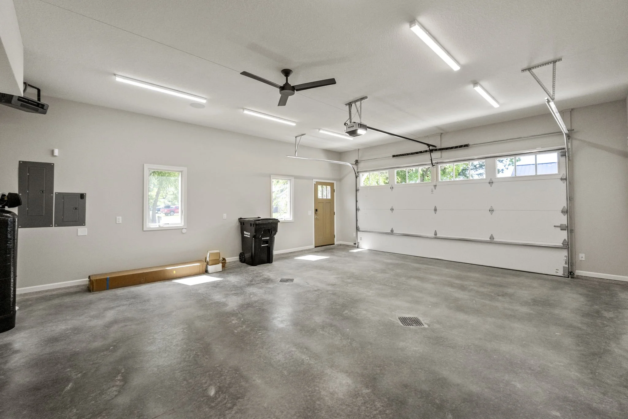 Empty garage with concrete floor, white walls, ceiling fan, fluorescent lights, three small windows, a closed garage door, a door to the house, and a few items including a waste bin, a long cardboard box, and a small box.