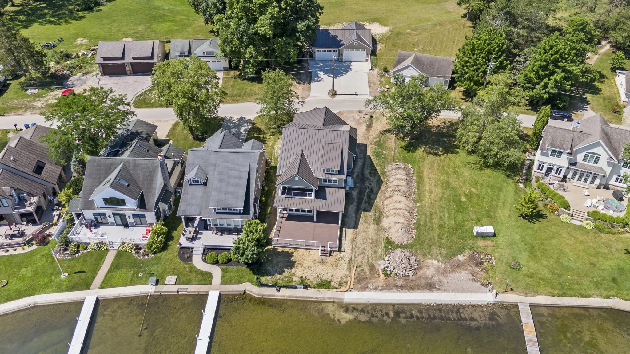 Aerial view of a neighborhood with multiple houses, trees, and a waterway with a dock. Some homes have decks, and there is a grassy area with construction or yard work happening.
