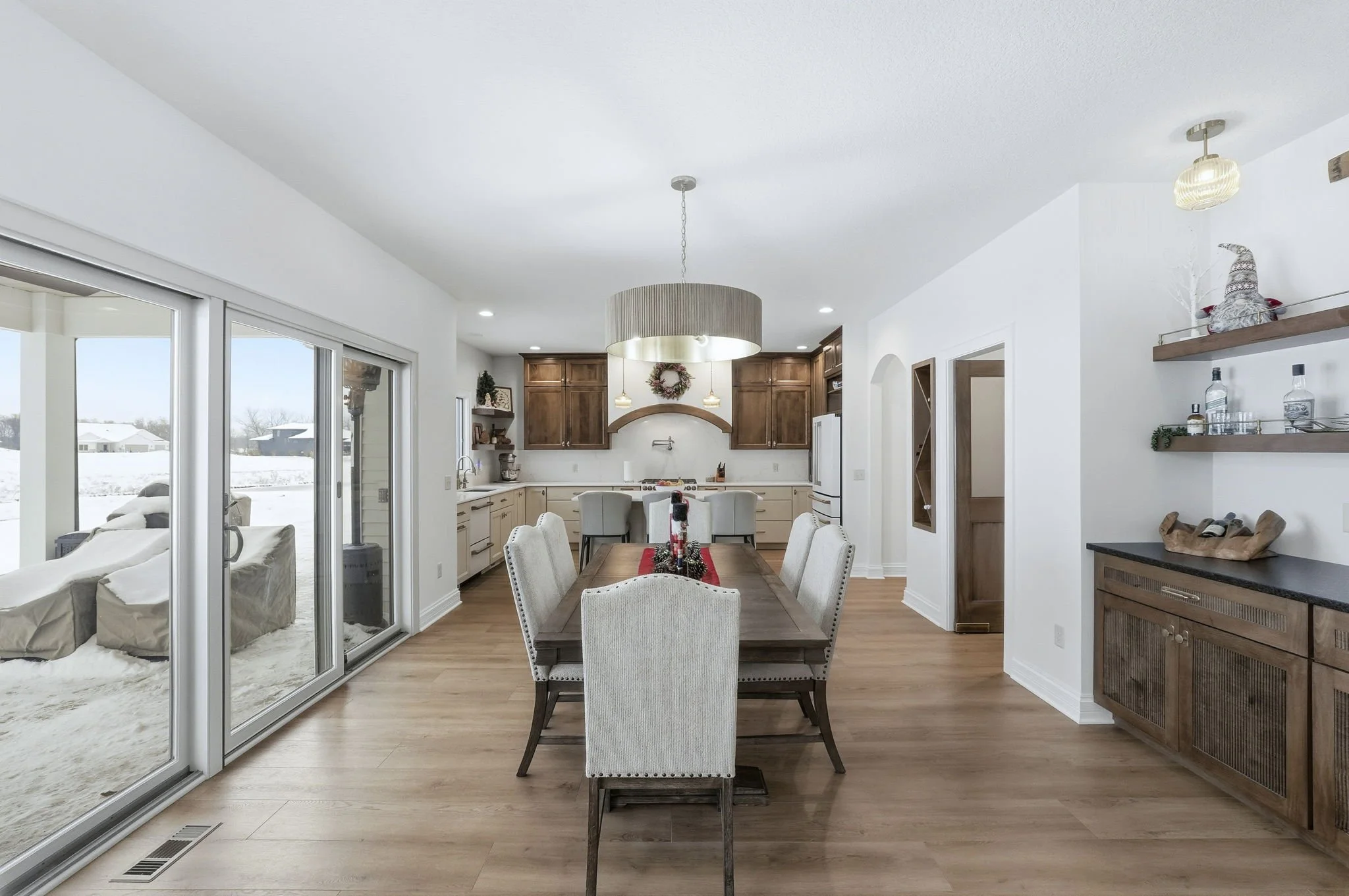 Open concept dining area and kitchen with white walls and light hardwood floors, decorated for Christmas with a wreath, table centerpiece, and holiday ornaments, adjacent to a sunroom with glass sliding doors revealing a snowy landscape outside.