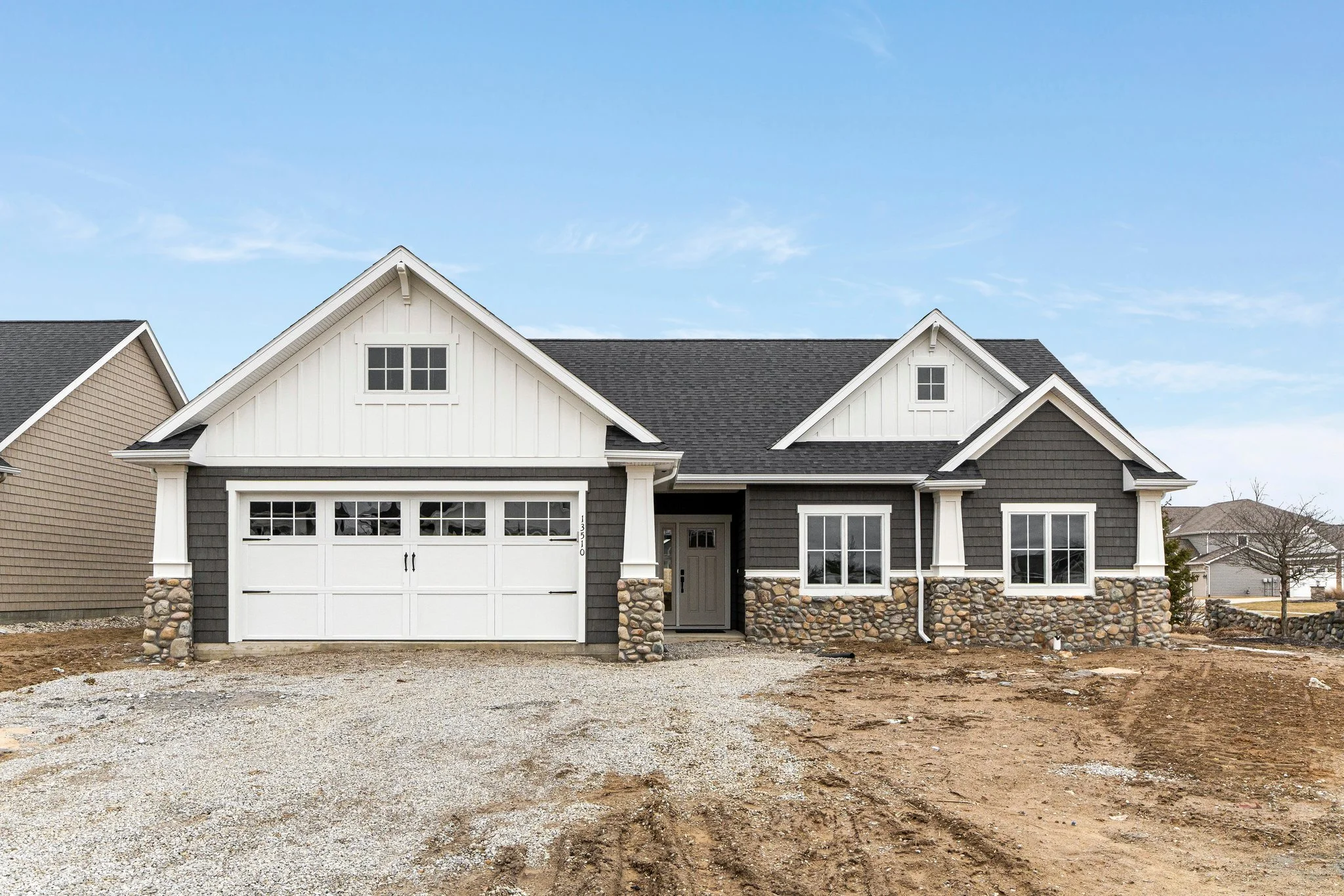 Newly constructed house with gray siding, stone foundation, white doors and window frames, and a gravel driveway in front.