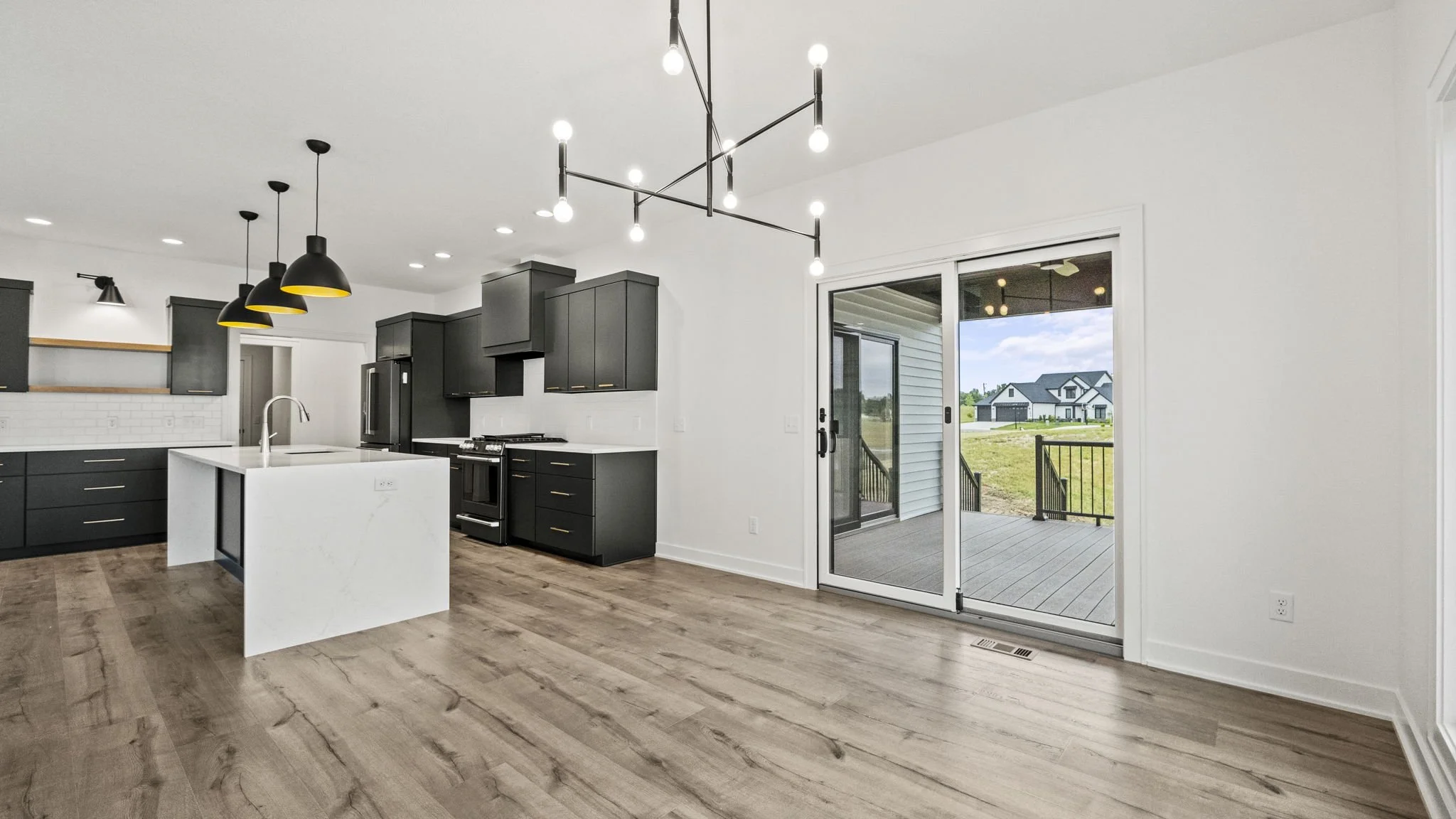 Modern kitchen with black cabinets, white island, and wood flooring, connected to a backyard deck through sliding glass door, with outdoor houses visible in the background.
