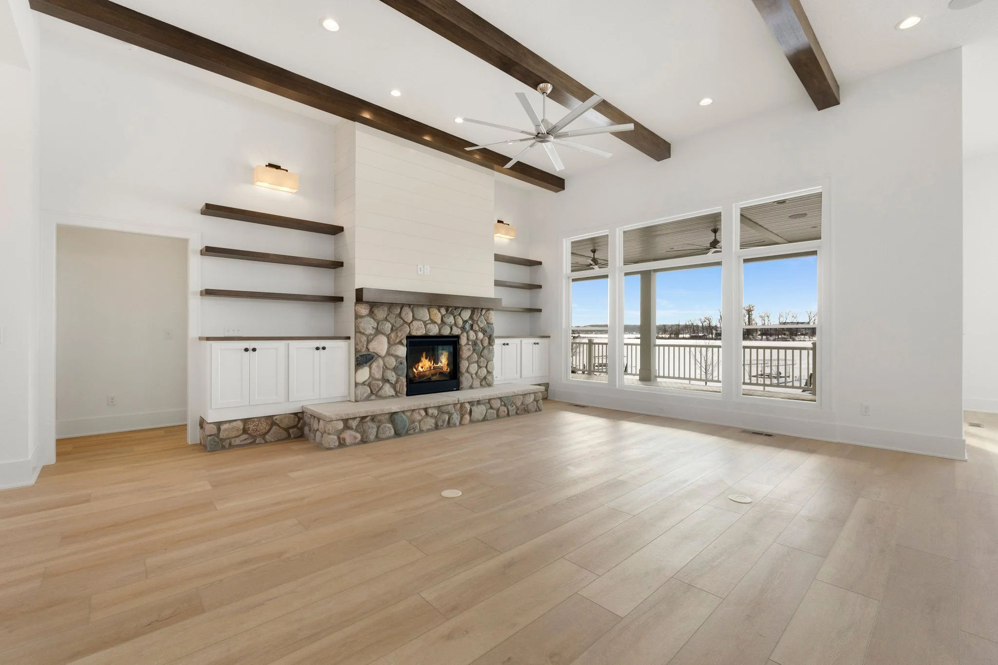 Empty living room with wooden floors, white walls, a stone fireplace, built-in white cabinets, and large windows showing a snowy outdoor landscape.