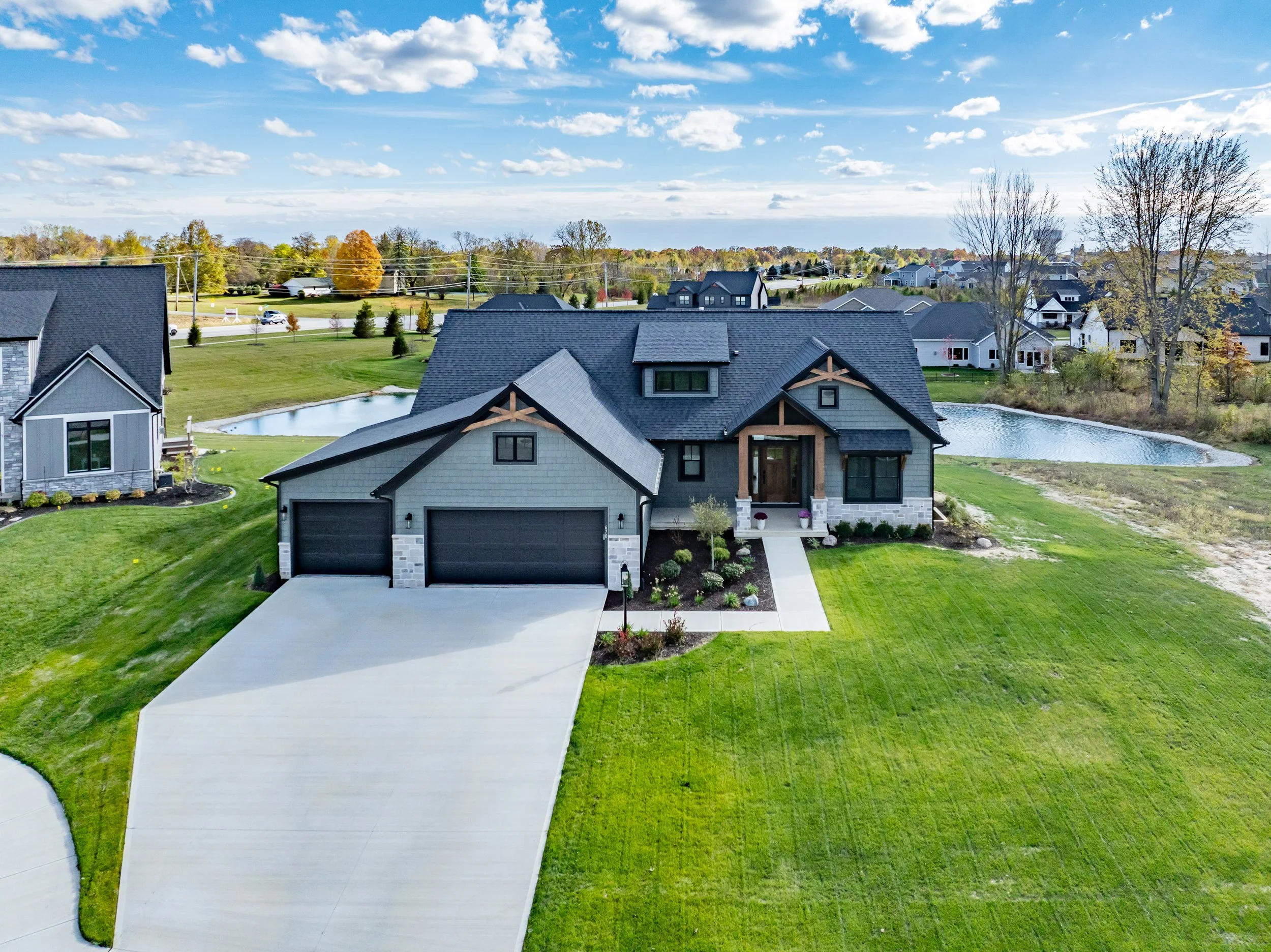 A modern two-story house with a dark shingle roof, gray siding, and stone accents, featuring a large driveway, manicured lawn, and surrounding pond, situated in a suburban neighborhood with other houses and trees in the background.