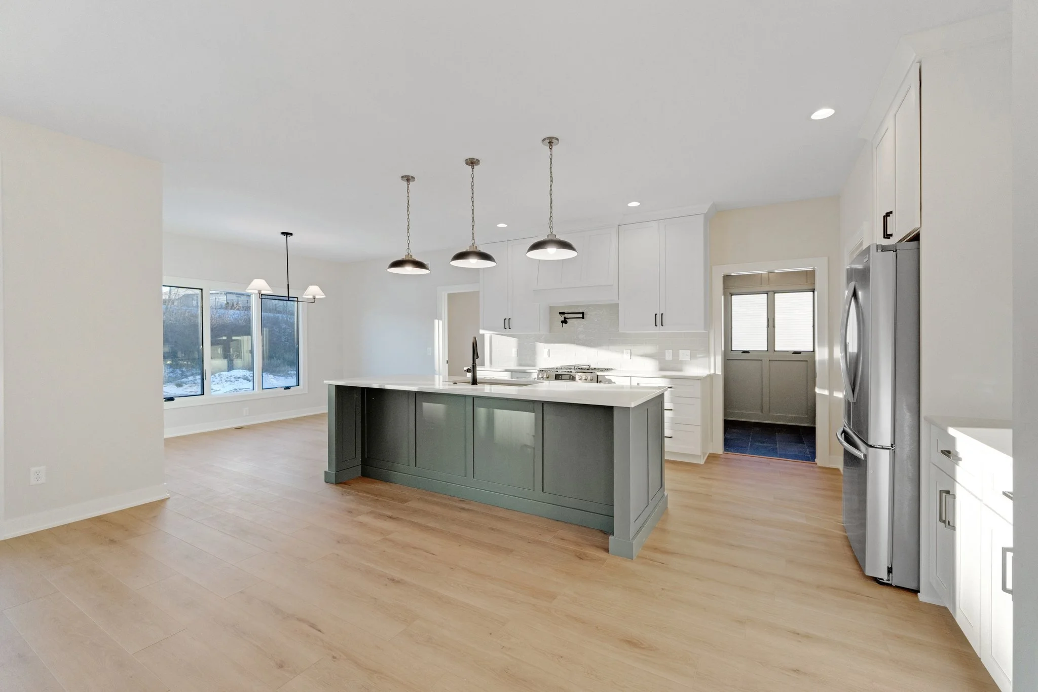 Open-concept kitchen with white cabinets, a dark gray island, stainless steel appliances, hardwood flooring, and large windows letting in natural light.