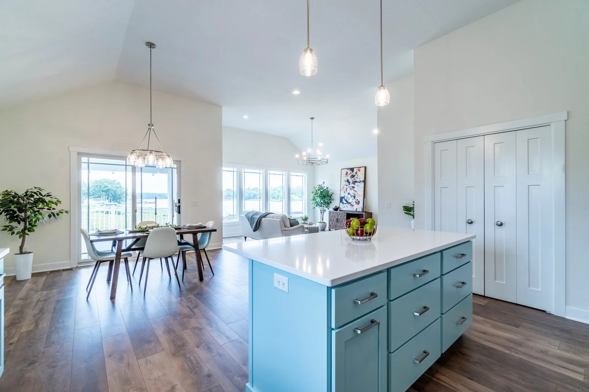 Open-concept living and dining area with a kitchen island, dining table, and sitting area, featuring large windows overlooking a waterfront view, with hardwood floors and modern decor.