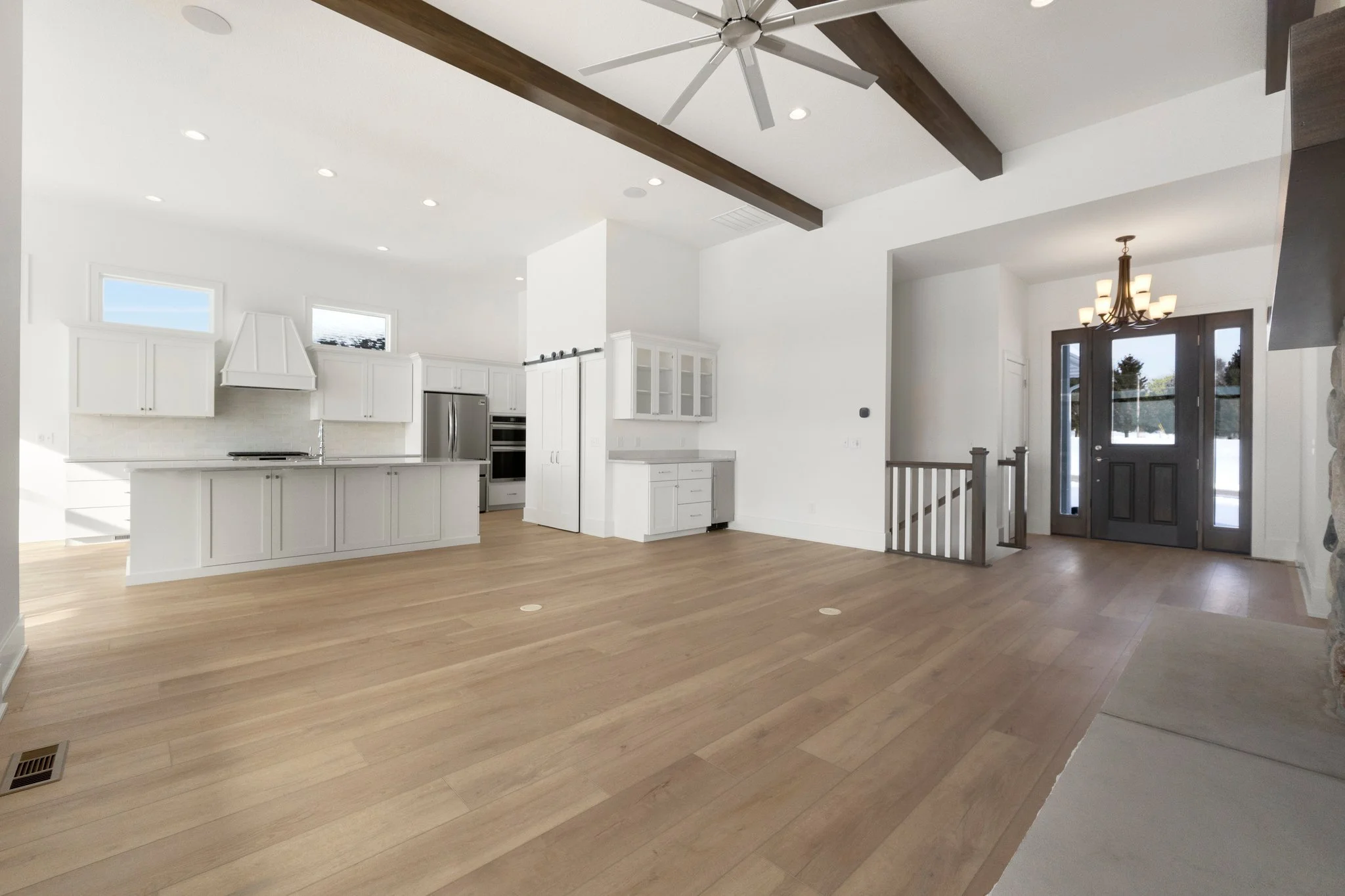 Spacious open-concept living area with white kitchen cabinets, stainless steel appliances, light wood flooring, exposed ceiling beams, and a front entry door with a chandelier overhead.