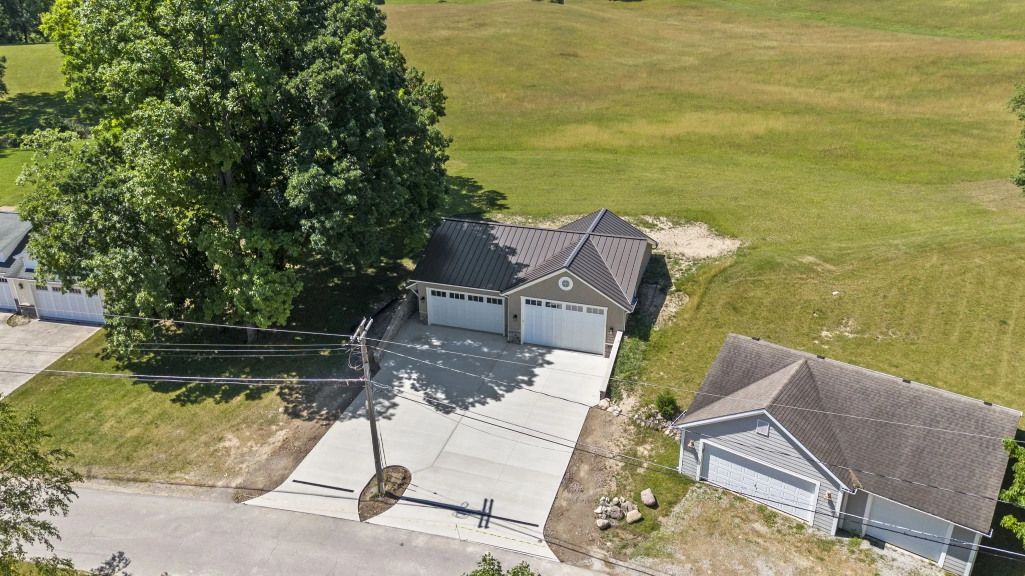 An aerial view of a residential area showing a house with a gray metal roof, a large driveway, and a big tree in the backyard, with open grassy fields surrounding the property.