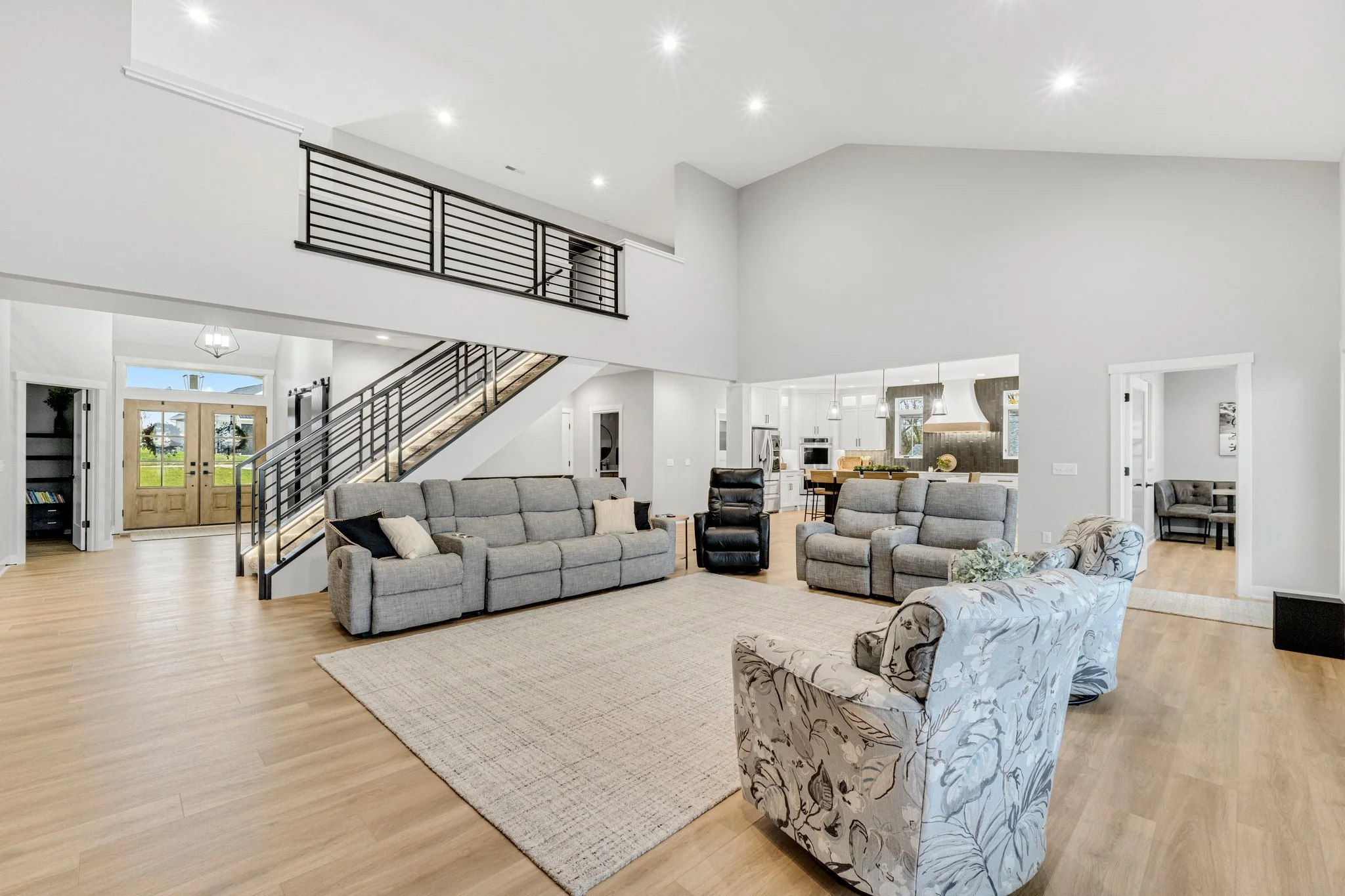 Spacious living room with gray sofas, floral armchair, area rug, open kitchen in background, light wood floors, white walls, black staircase railing.
